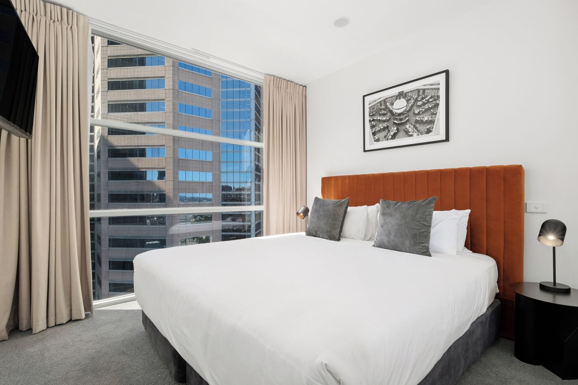 Een hotelkamer met een groot bed, hoofdbord, een inlijsting foto aan de muur, een raam met gordijnen en een nachtkastje met een lamp.