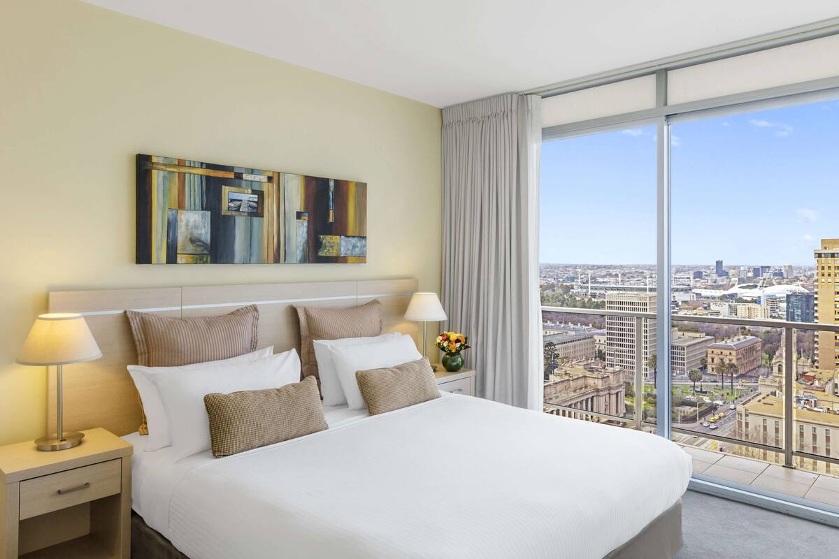 A hotel room with a large bed, a nightstand, a lamp, a vase with flowers, and a city view through large windows.