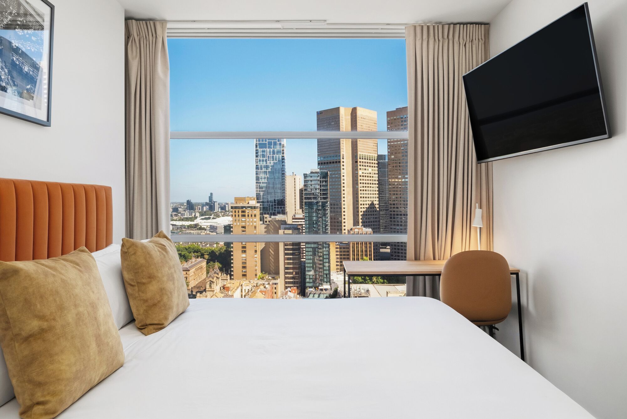 Quarto de hotel com uma janela grande que oferece vista da cidade, uma cama com duas almofadas, uma mesa com uma cadeira e uma TV de tela plana fixada na parede.