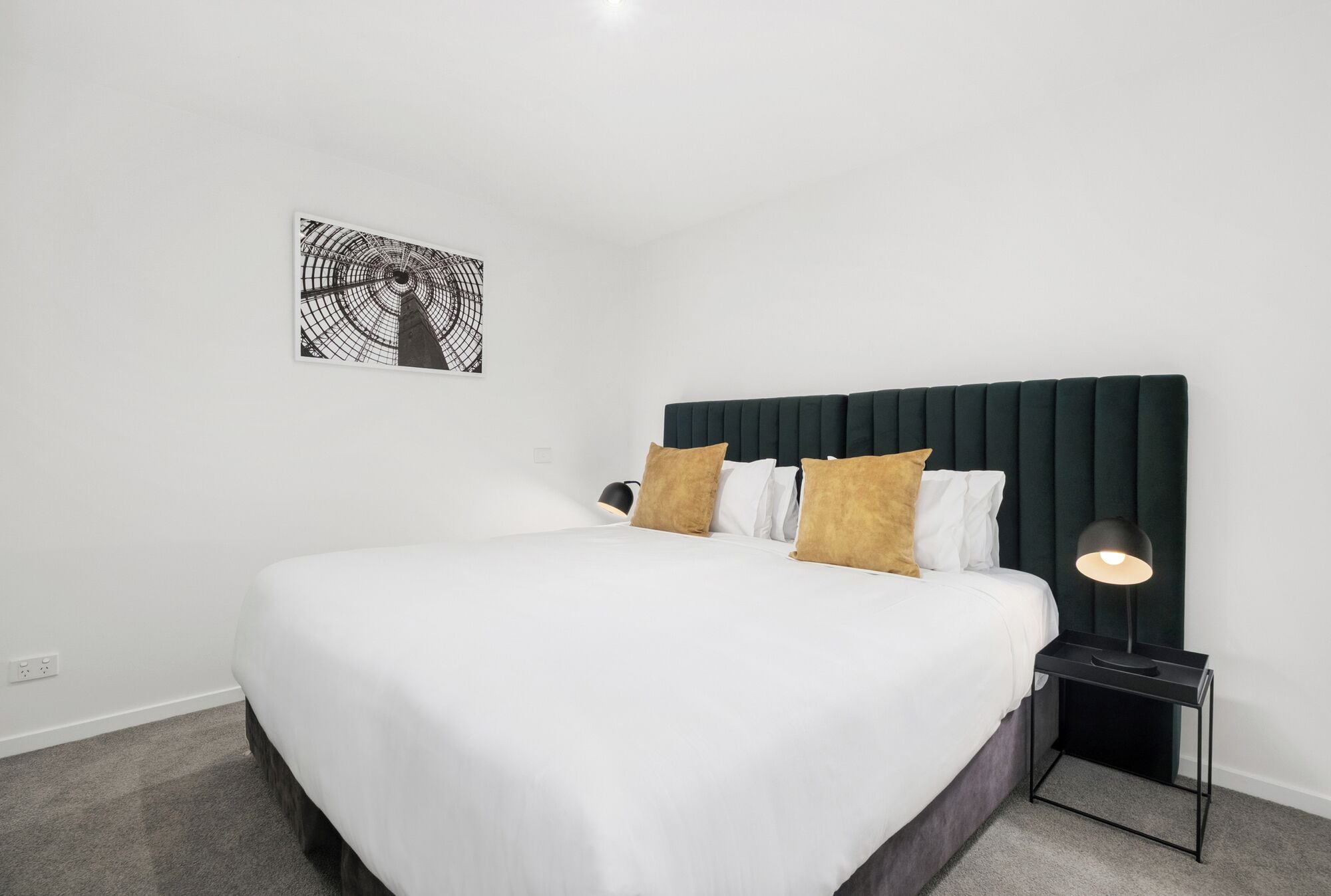 A neatly made bed with white bedding and two brown pillows, a dark green upholstered headboard, a black side table with a lamp, and a black and white abstract artwork on the wall.