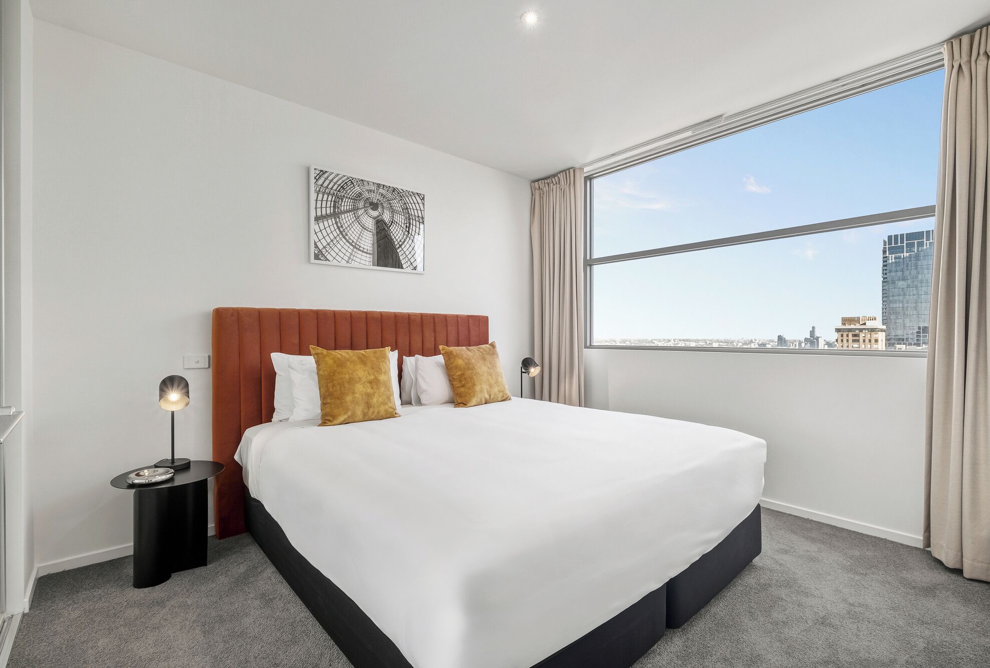 Quarto de hotel moderno com uma cama grande, vista para a cidade e uma obra de arte emoldurada na parede.
