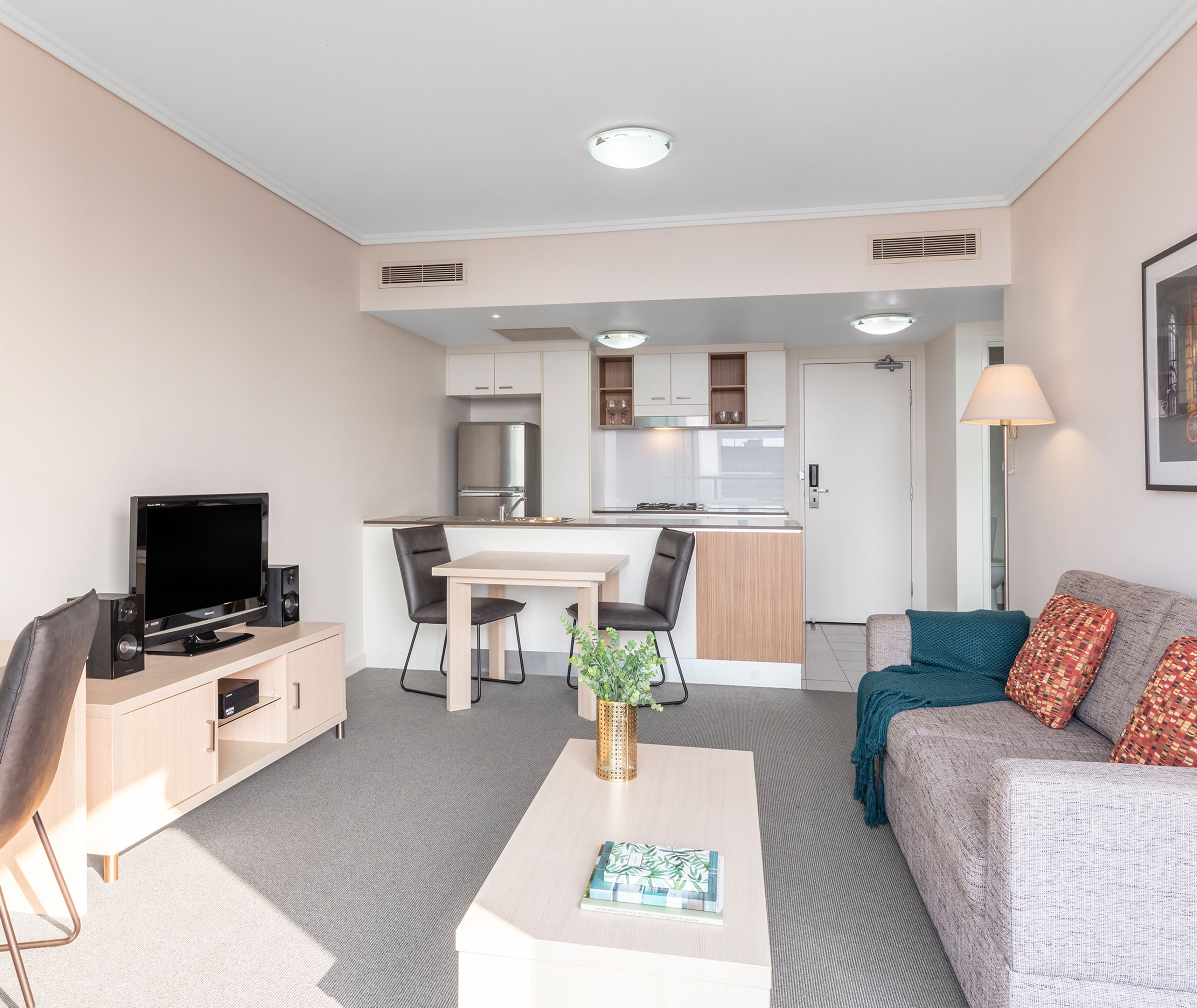 Ein modernes Wohnzimmer mit Flachbildfernseher, Couchtisch mit Buch, Sofa mit gemusterten Kissen und einer Küchenzeile mit Esstisch und Stühlen.