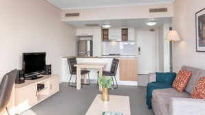 Una sala de estar moderna con un televisor de pantalla plana, una mesa de centro con un libro, un sofá con cojines estampados y una zona de cocina con una mesa de comedor y sillas.