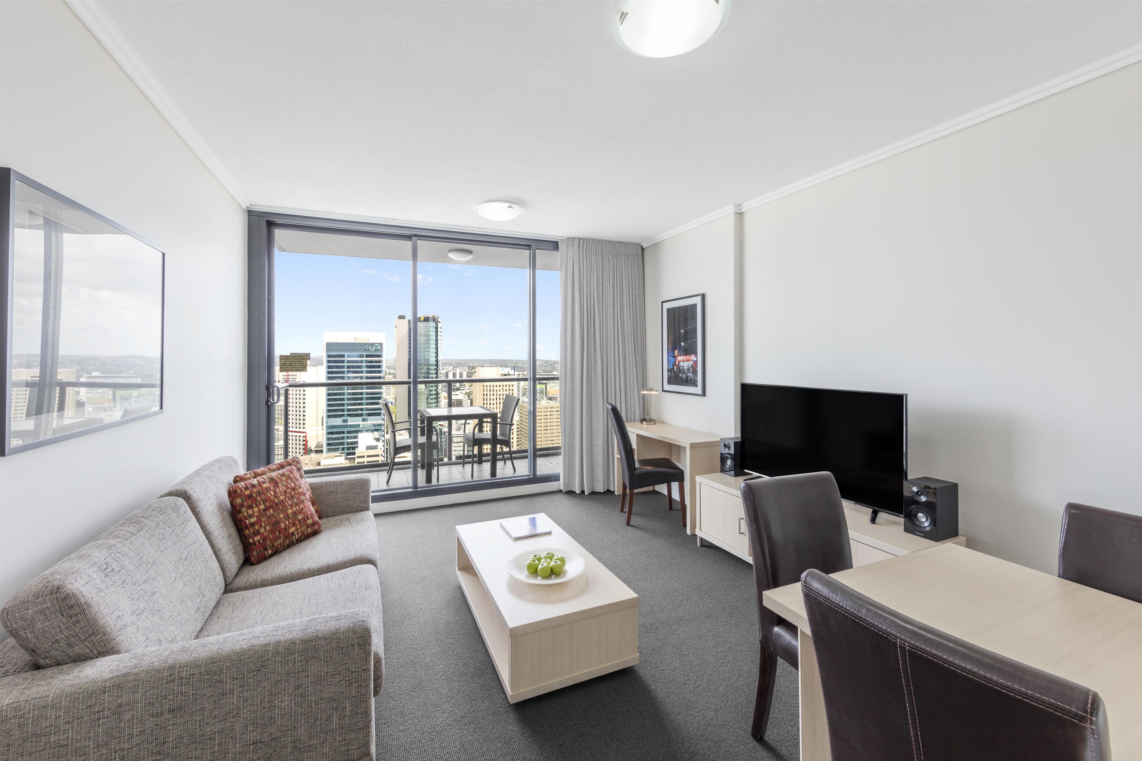 A modern hotel room with a balcony, a sofa, a dining table, a TV, and a desk.
