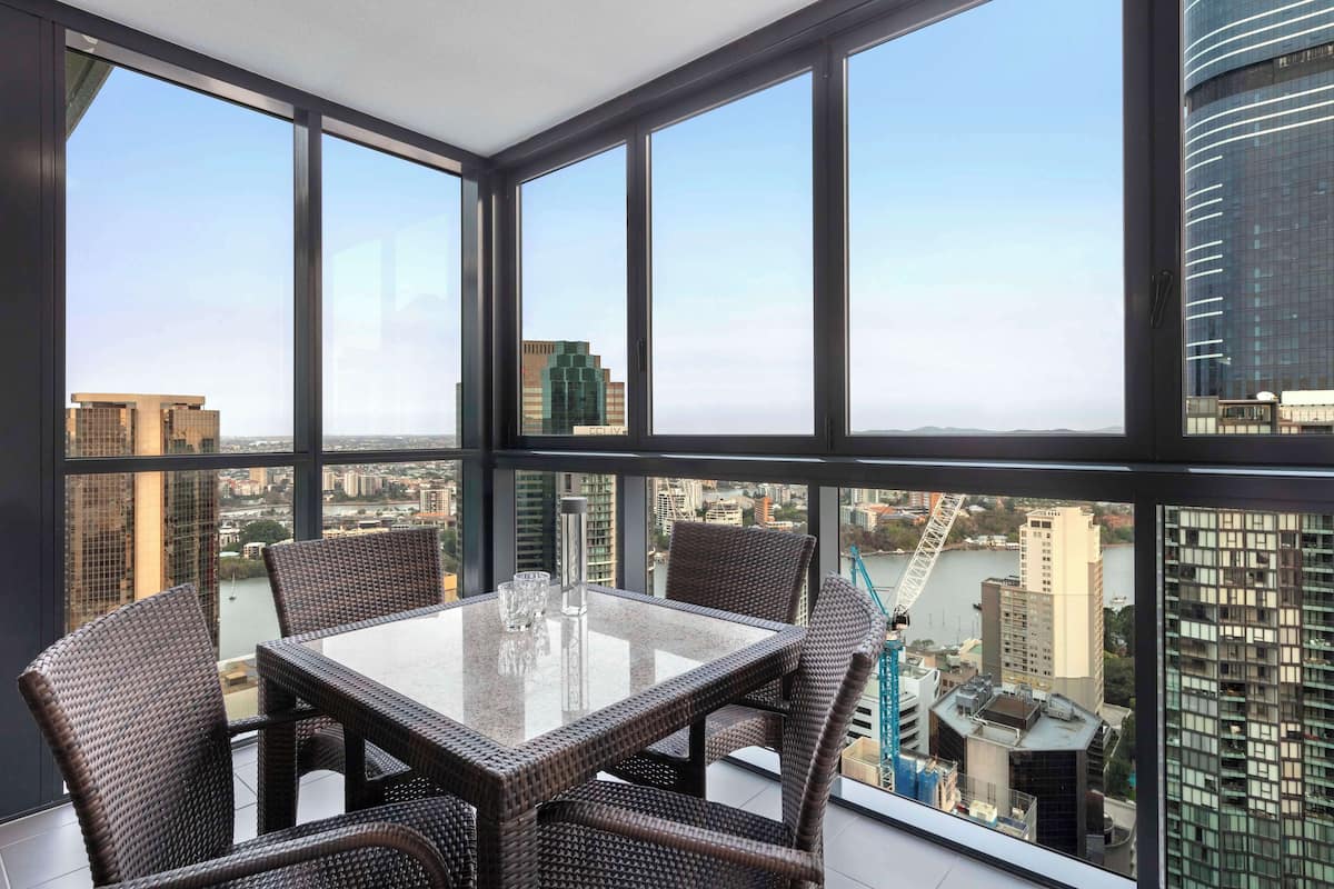 Een hoog gelegen kamer met een groot raam met uitzicht op een stadsgezicht, een tafel met glazen blad en rieten stoelen.