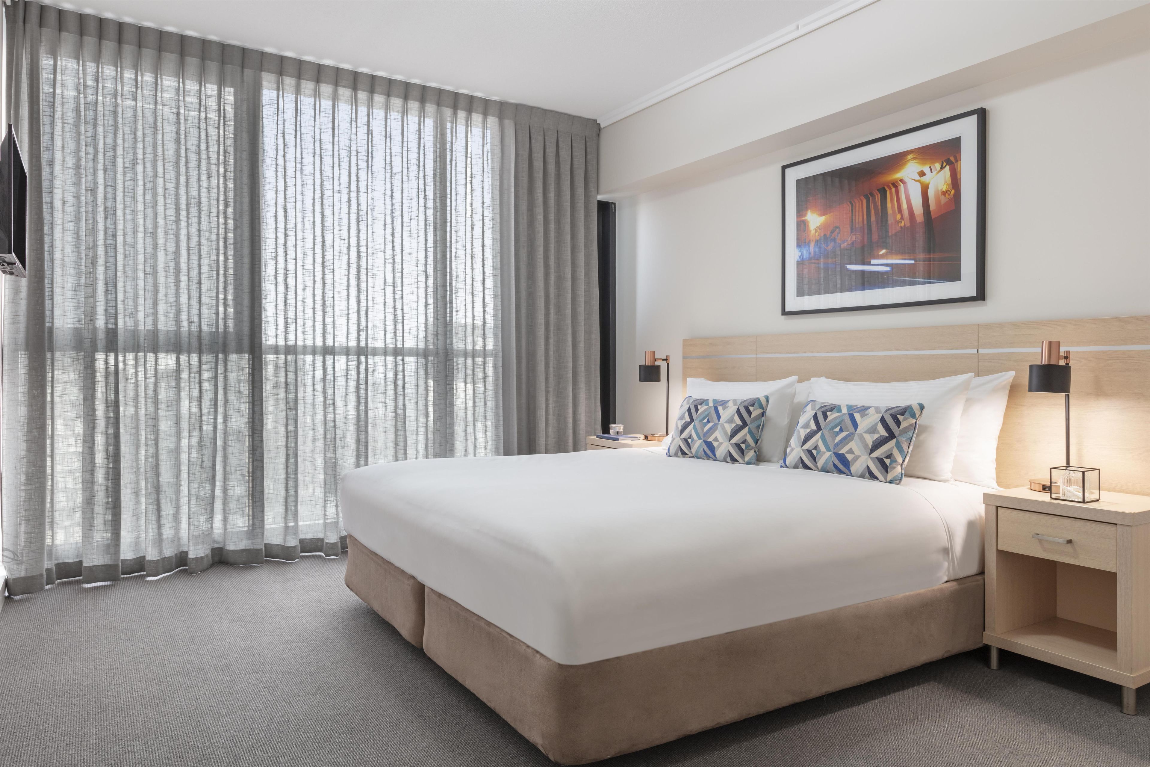 A hotel room with a large bed, a nightstand, a lamp, a framed picture on the wall, and sheer curtains.