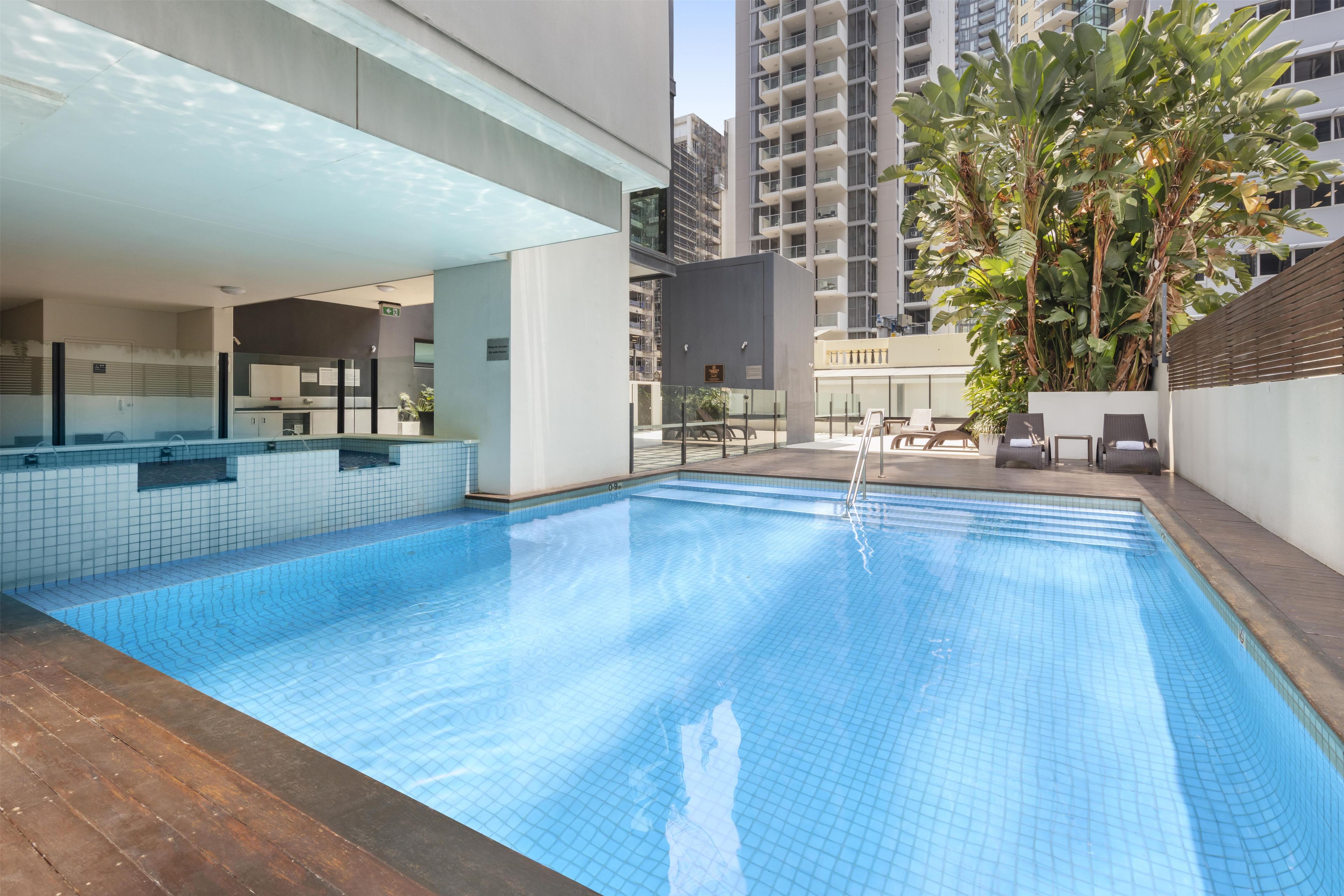 Piscina com deck revestido, cercada por um edifício com design moderno, incluindo varanda e área coberta.