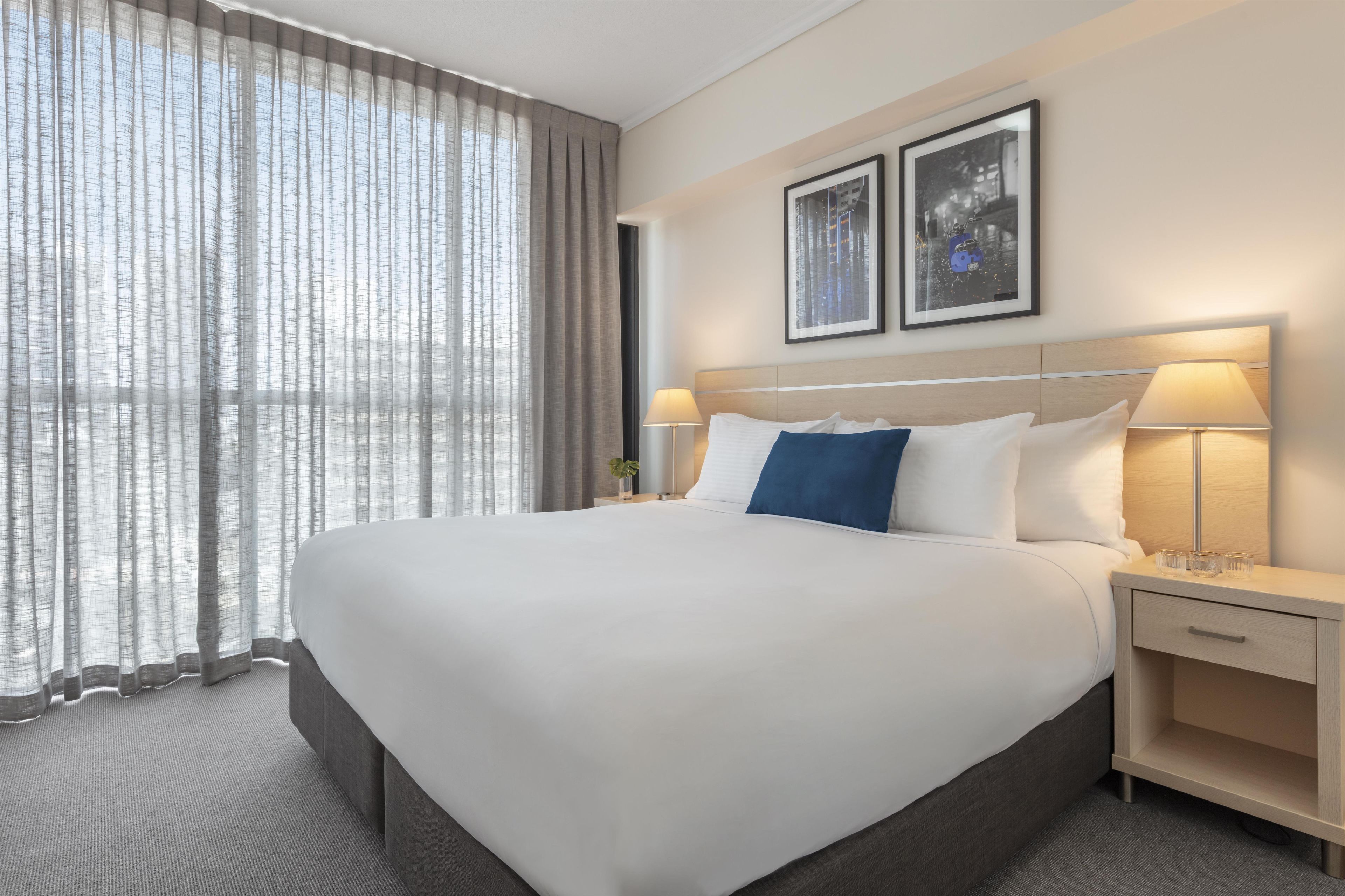Habitación de hotel con una cama grande, dos lámparas de noche, una mesita de noche y una ventana con cortinas traslúcidas.