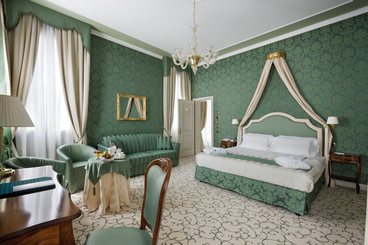 Une chambre à coucher avec un lit à baldaquin, un fauteuil vert, un bureau en bois et un lustre.