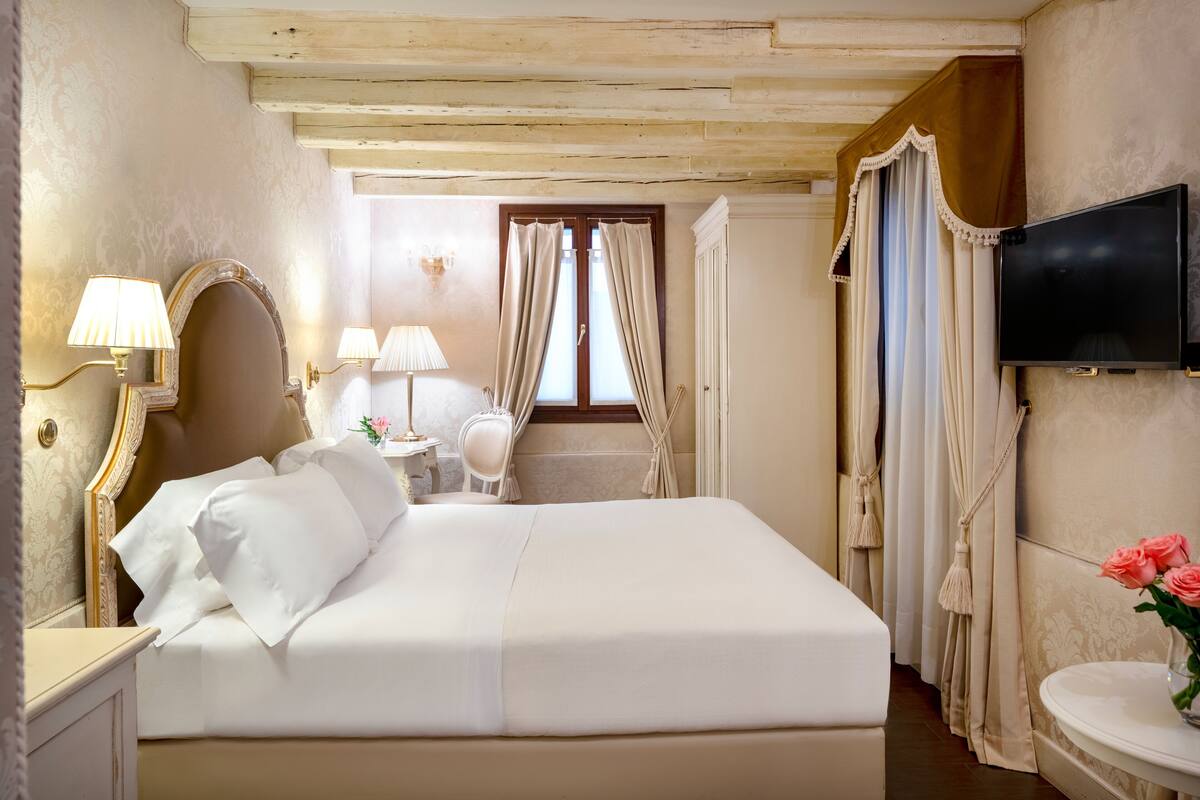 Ein Hotelzimmer mit einem großen Bett, einem an der Wand befestigten Fernseher und einem kleinen Tisch mit Blumenvase.