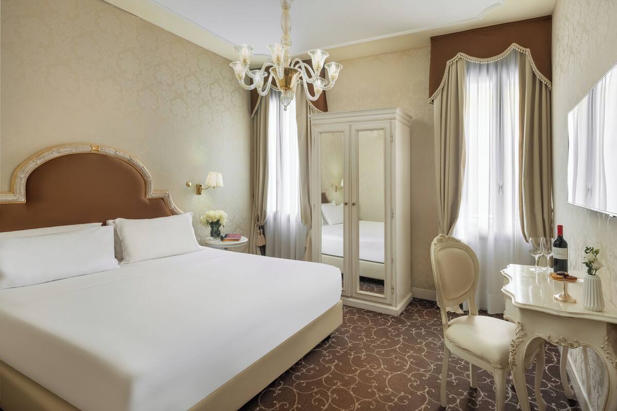 Ein Hotelzimmer mit einem großen Bett, einem Schminktisch mit Spiegel, einem Sessel und einer Kristallleuchter.