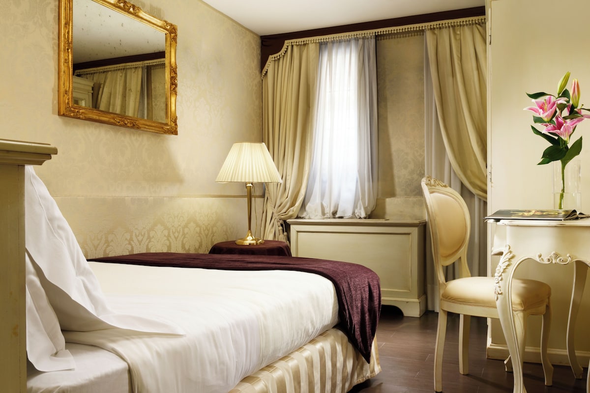 Une chambre d’hôtel avec un lit, un bureau, une chaise, une lampe et un miroir.
