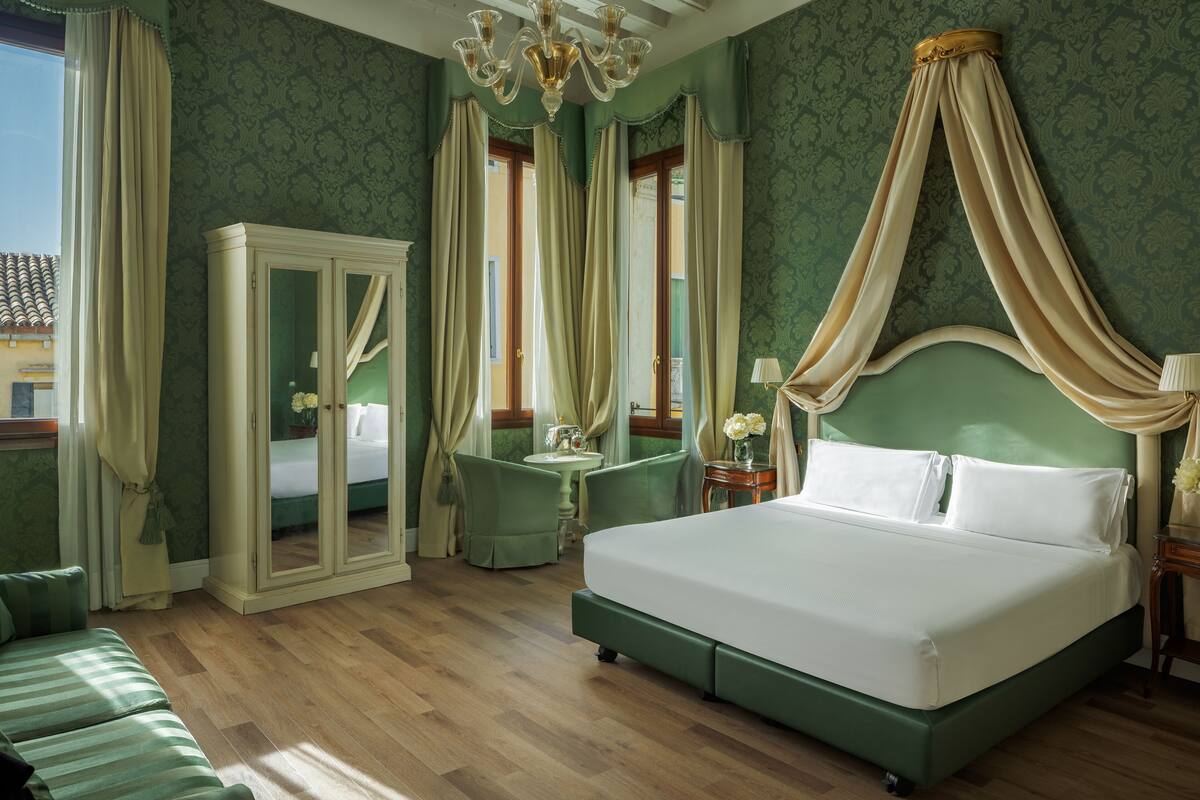 Ein Schlafzimmer mit einem großen Bett, einem Schminktisch mit Spiegel, einem grünen Sessel und einem Fenster mit Vorhängen.