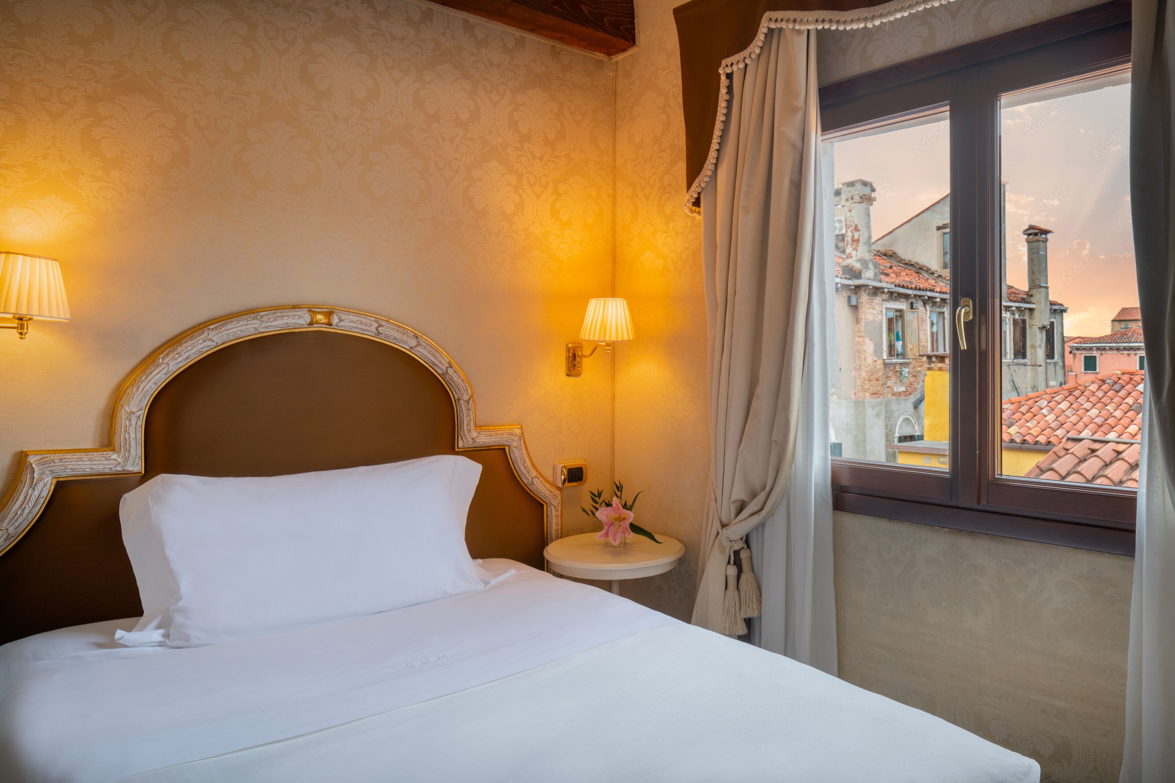 Une chambre d’hôtel avec un lit, deux lampes fixées au mur, une table ronde ornée d’un vase de fleurs et une fenêtre avec des rideaux.