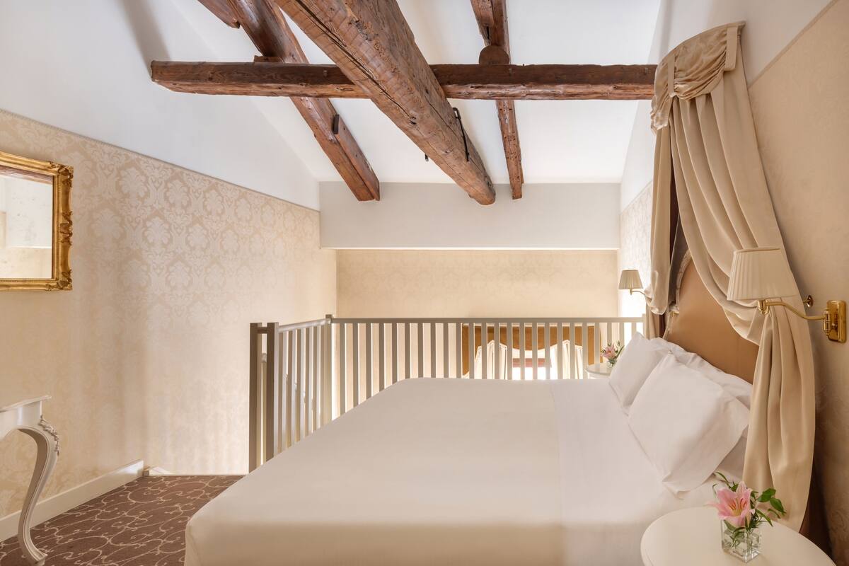 Ein Schlafzimmer mit einem großen Bett, Holzbalken, einem weißen Kopfteil, einem Nachttisch mit einer Vase und einem Spiegel an der Wand.