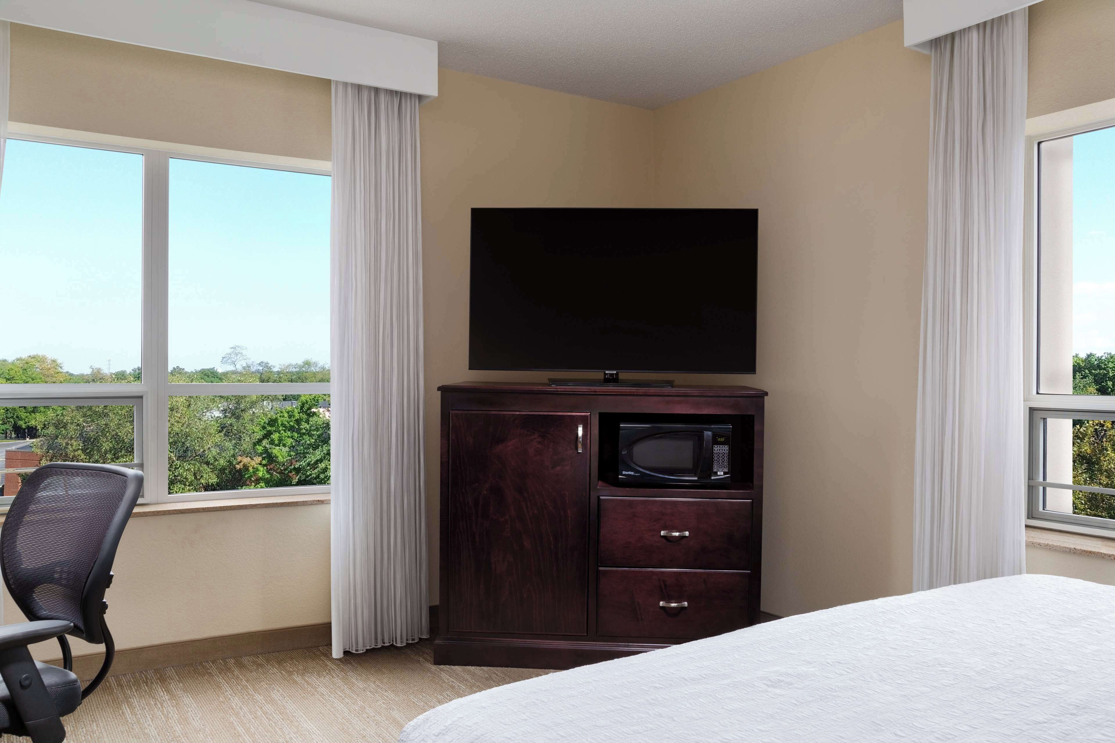 A hotel room with a bed, a TV on a wooden cabinet, a microwave, a chair, and large windows offering a view of greenery.