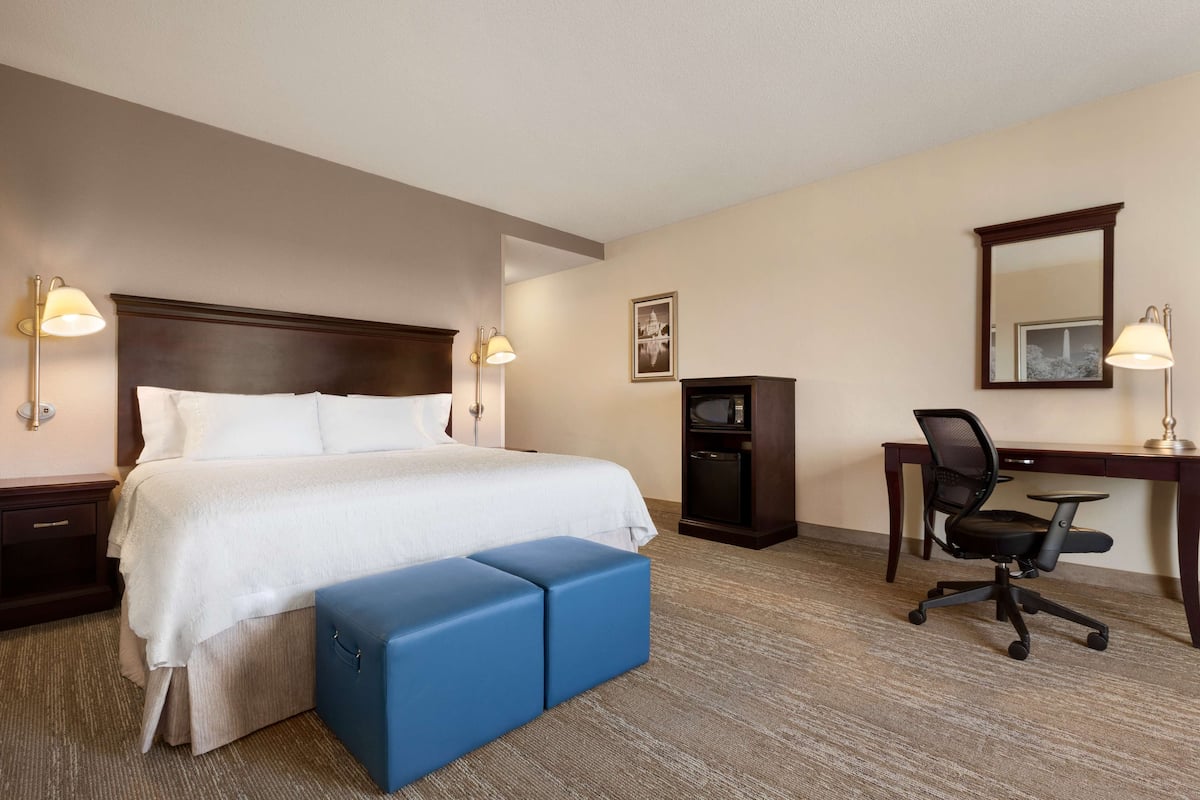A hotel room with a large bed, a desk, a chair, and a small blue ottoman.