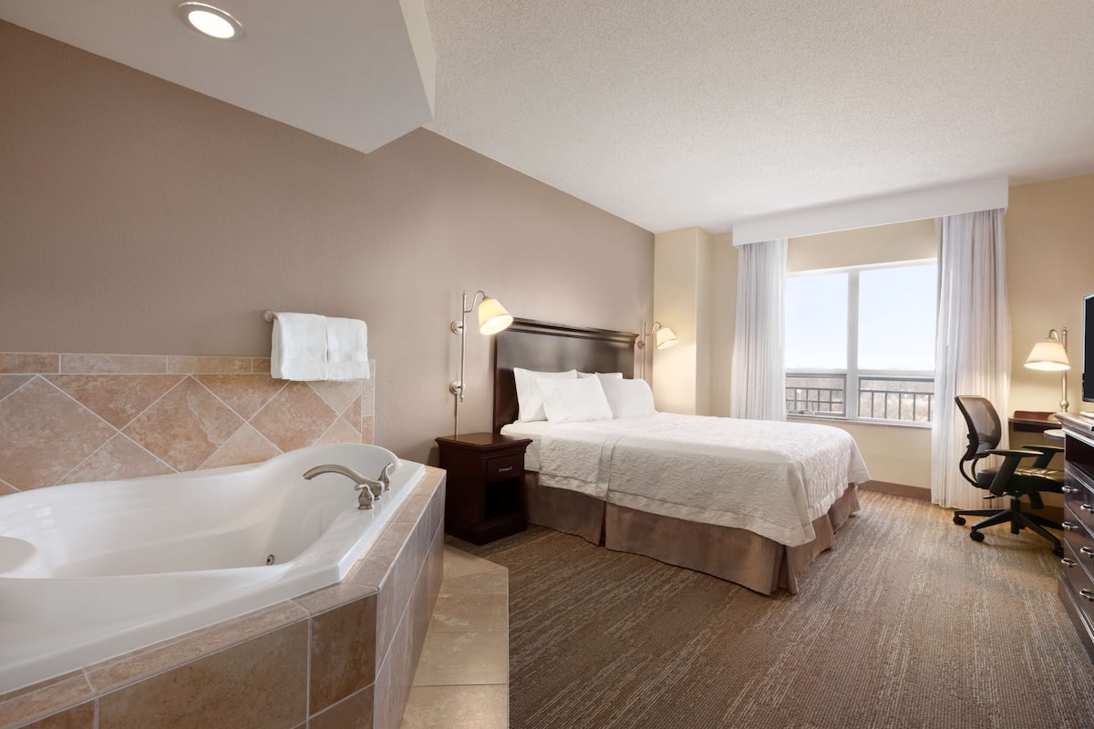 A hotel room with a large bathtub, a bed with a headboard, a desk with a chair, and a window with curtains.