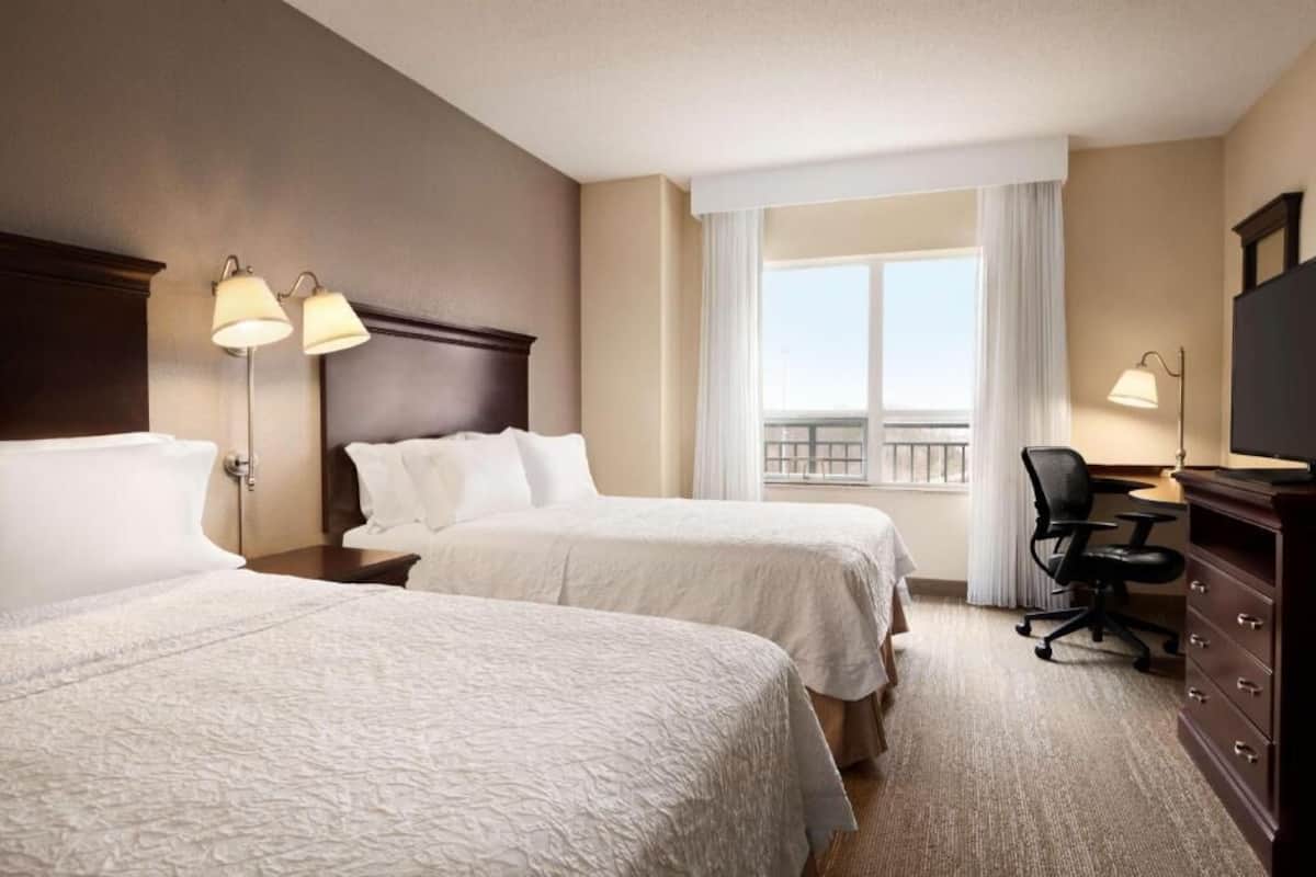 Habitación de hotel con dos camas, un escritorio, una silla, un aparador y un balcón con cortinas.
