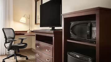 A hotel room with a desk, a computer monitor, a microwave, a trash bin, and a chair.