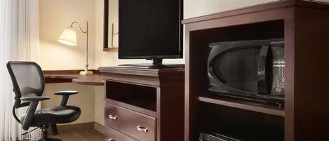 A hotel room with a desk, a computer monitor, a microwave, a trash bin, and a chair.
