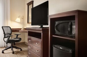 A hotel room with a desk, a computer monitor, a microwave, a trash bin, and a chair.