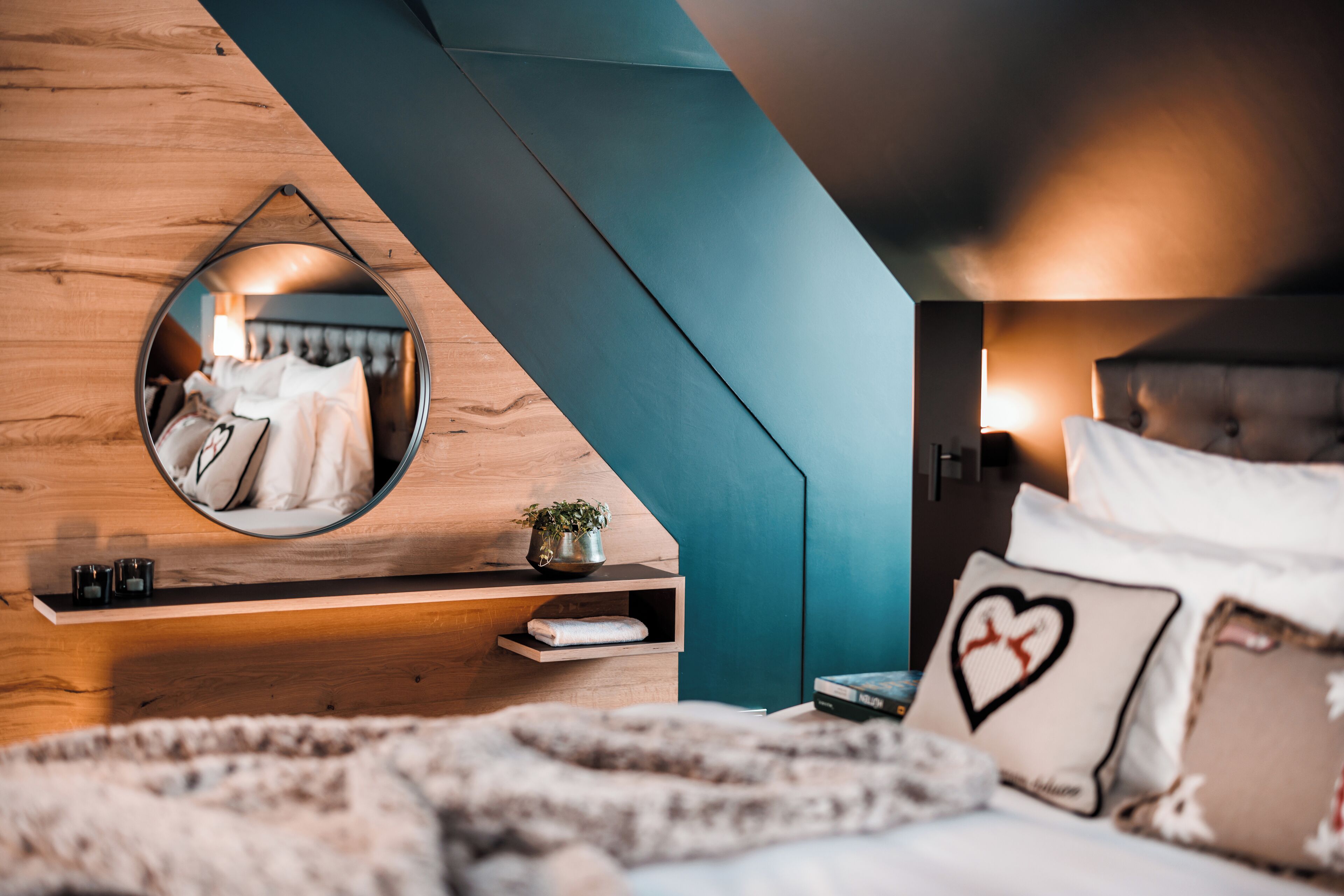 A bedroom with a wooden headboard, a circular mirror, a teal accent wall, and a bed with a fluffy blanket and heart-shaped pillow.