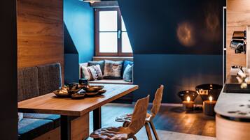 A cozy dining area with a wooden table, two chairs, and a bench. There are candles on the floor and a window with daylight coming through.