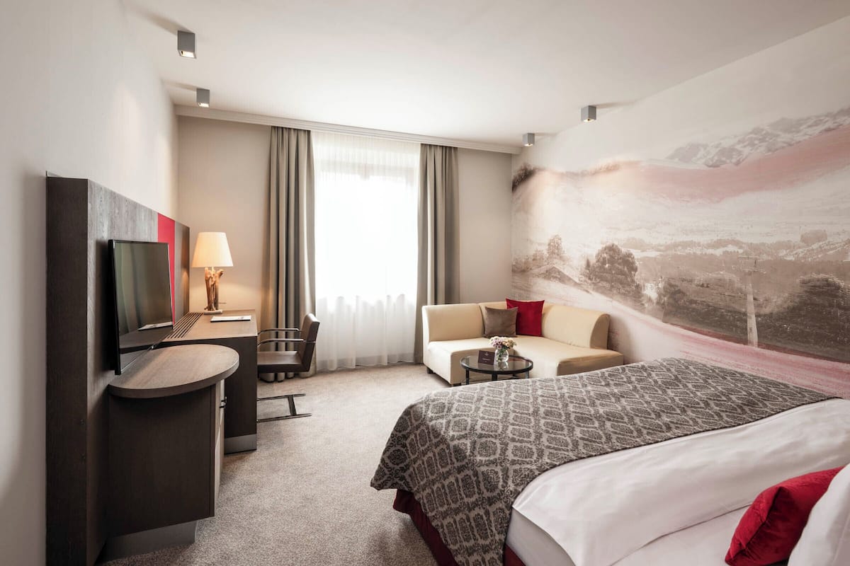 Ein Hotelzimmer mit einem Bett, einem Schreibtisch mit Stuhl, einer Couch, einem Fernseher und einem großen Fenster mit Vorhängen.