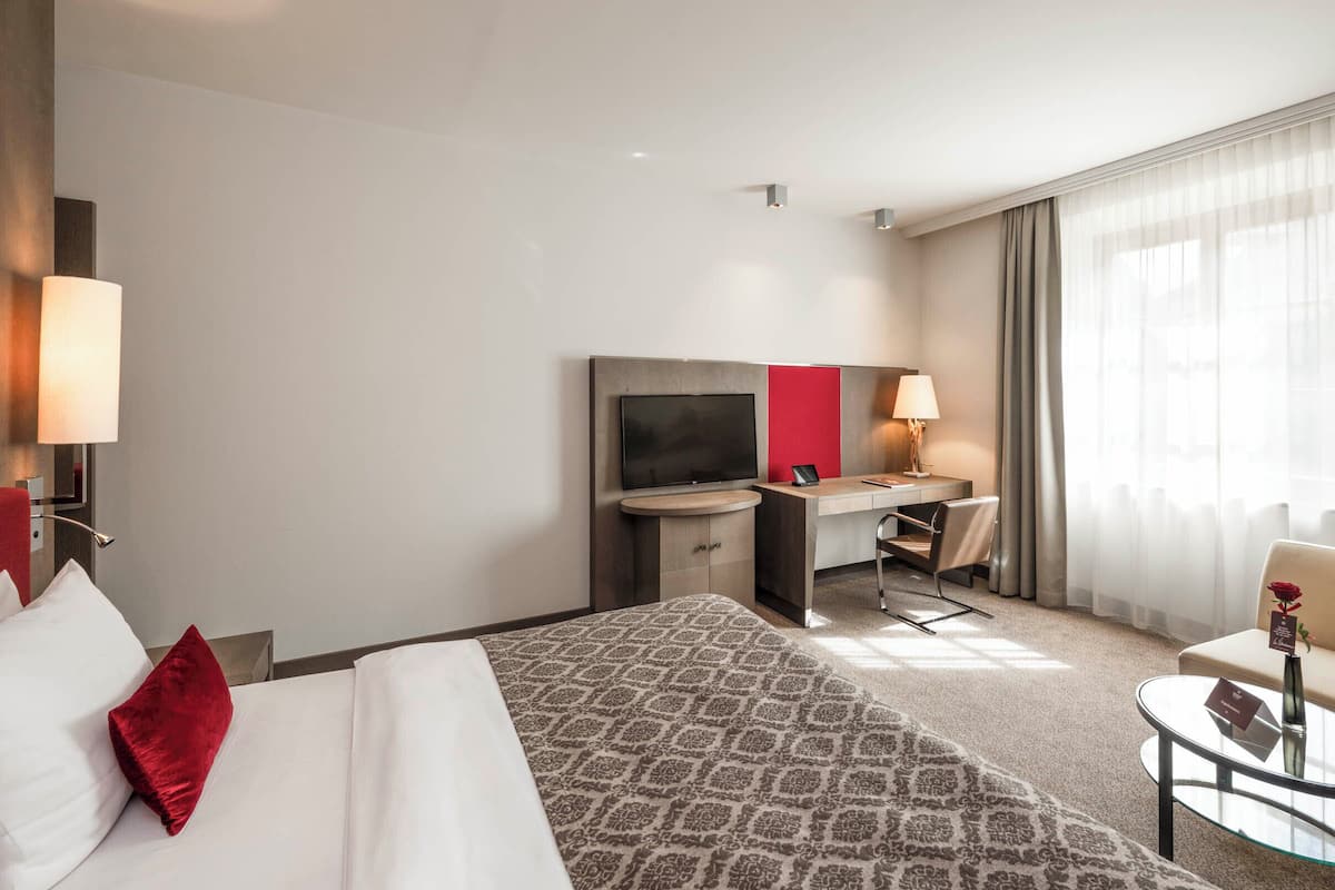 Ein Hotelzimmer mit Bett, Schreibtisch mit Fernseher, Stuhl, kleinem Tisch und einem Fenster mit Vorhängen.