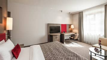 Habitación de hotel con cama, escritorio con televisor, silla, mesita y ventana con cortinas.