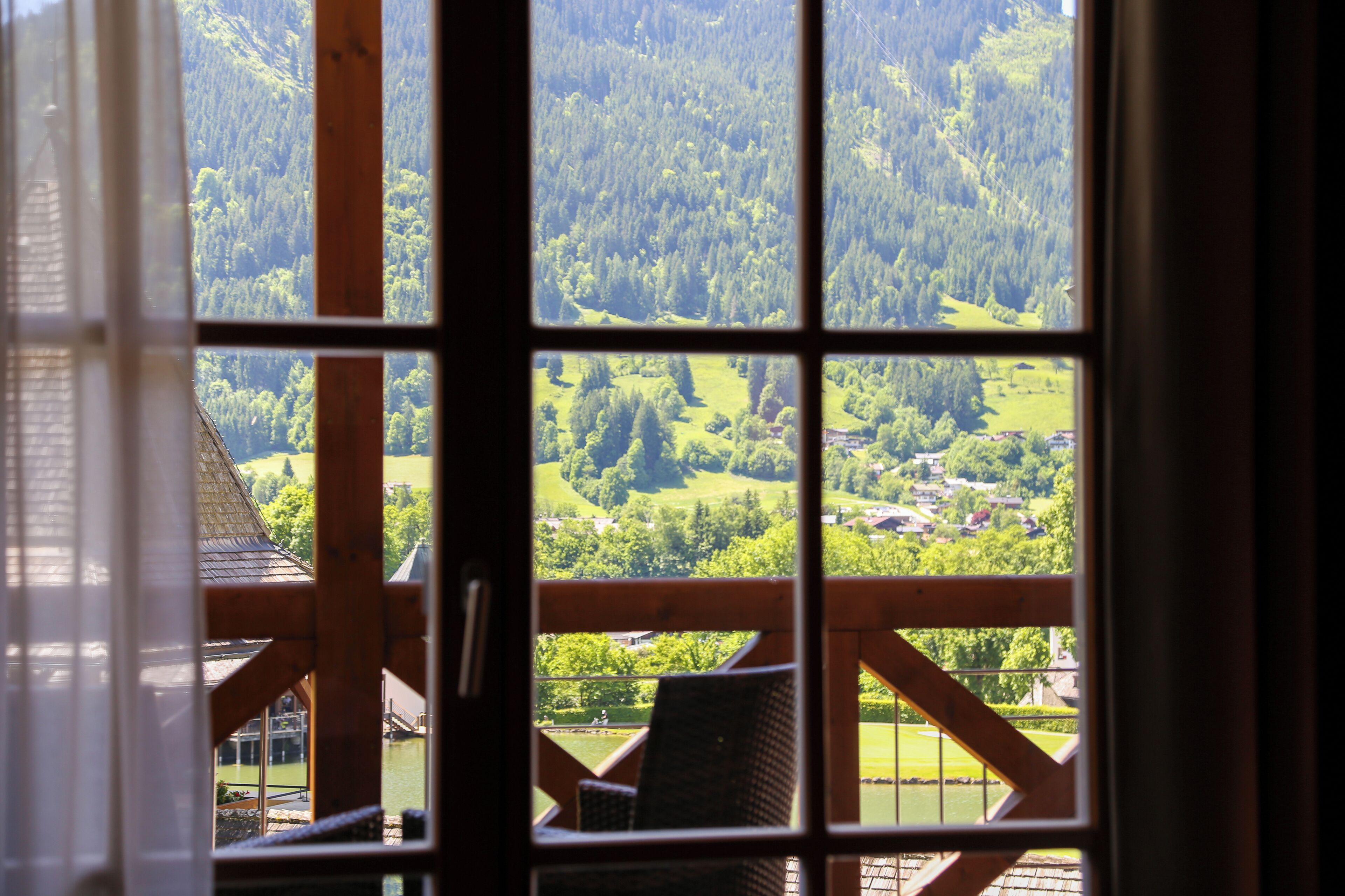 A view from a window with a wooden frame, overlooking a green landscape with trees and a small village.