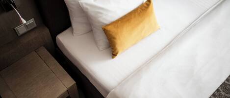 A neatly made bed with white linens and a mustard-colored pillow, a wooden nightstand, and a wall-mounted lamp.