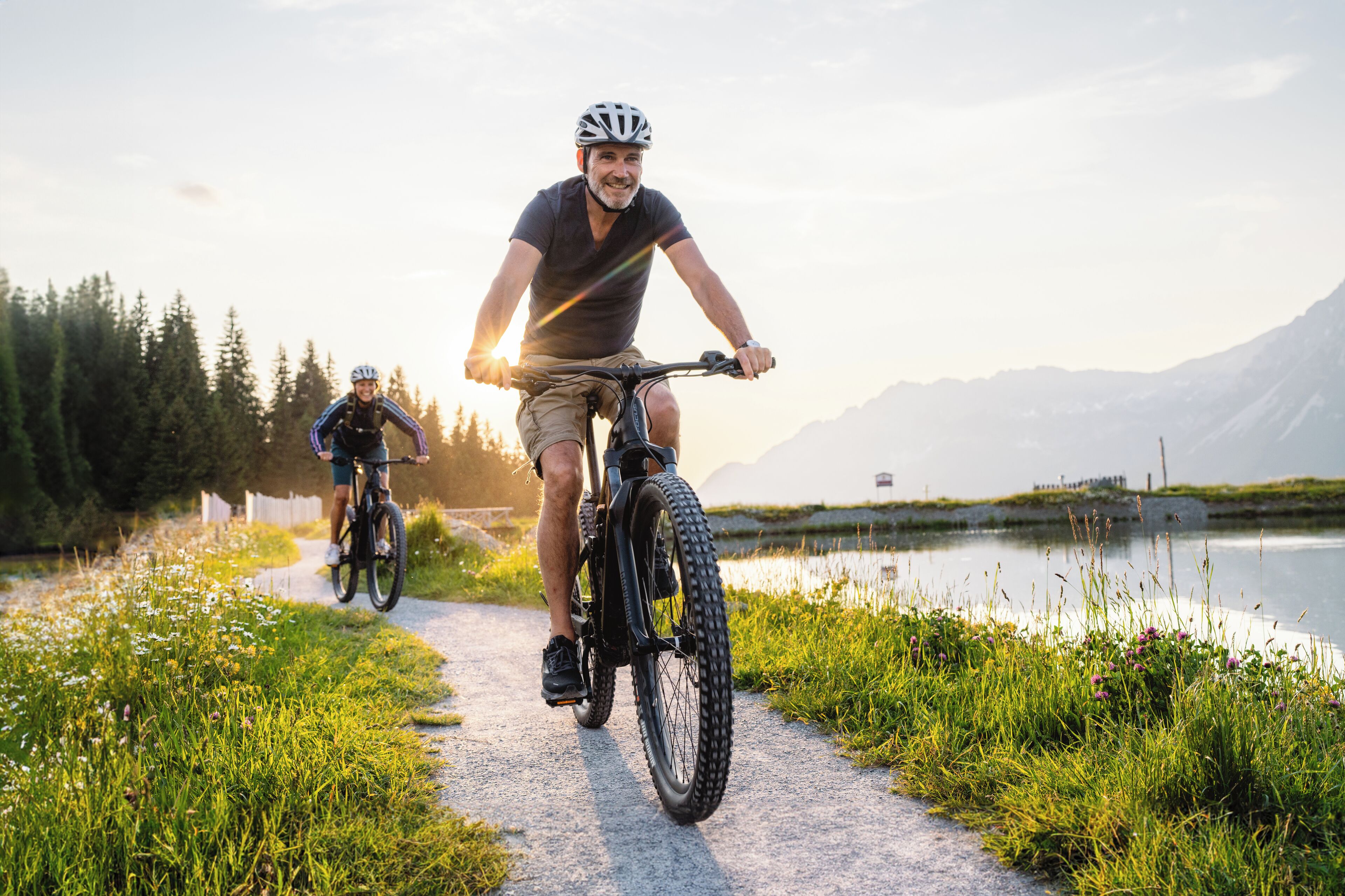 Two cyclists on a paved path beside a lake with mountains in the background.