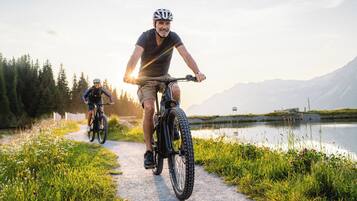 Two cyclists on a paved path beside a lake with mountains in the background.