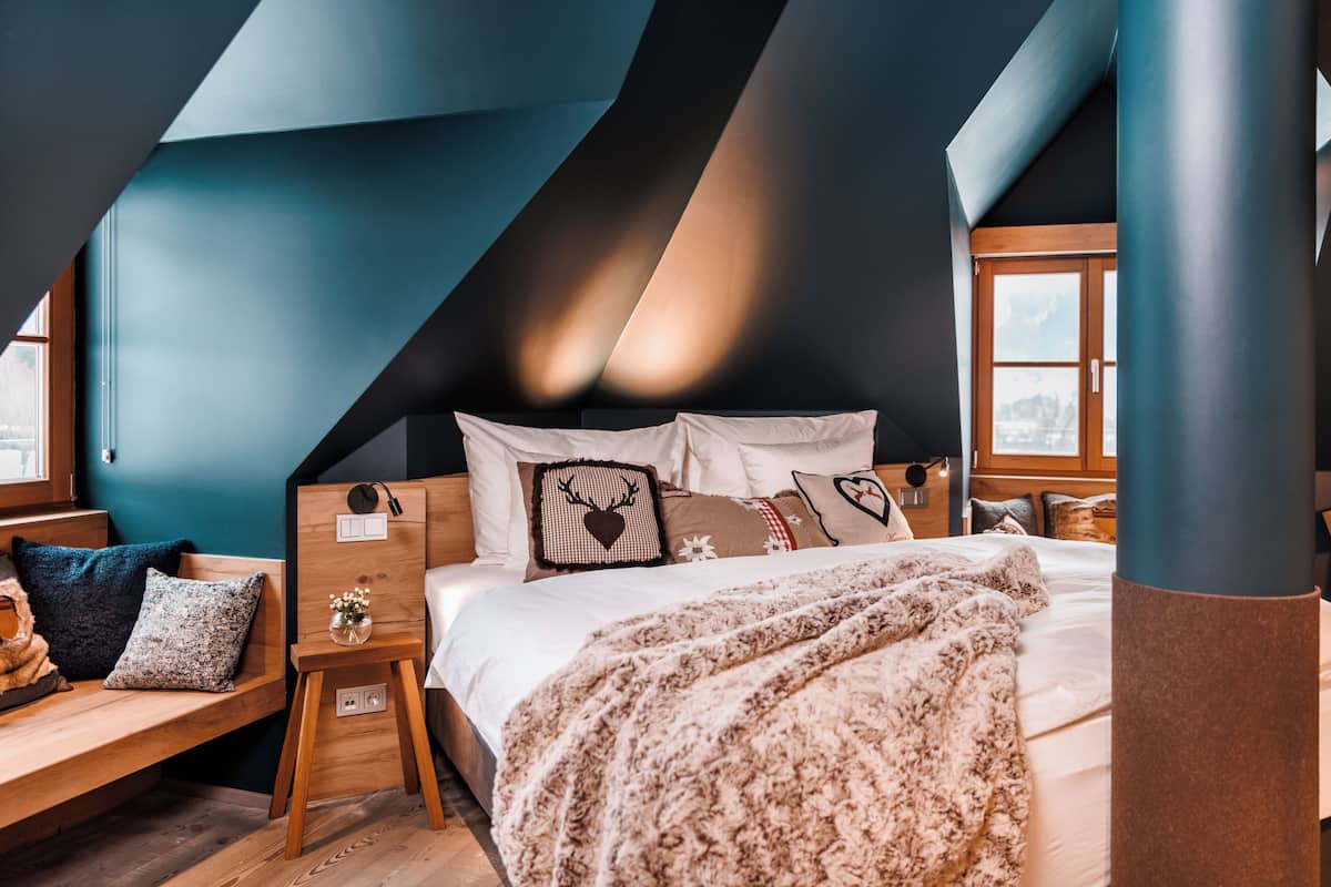 Ein Schlafzimmer mit schrägen Wänden, einem Bett mit vielen Kissen, einem Nachttisch mit Lampe und einem Fenster mit Ausblick.