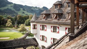 A traditional European-style building with multiple windows and a sloped roof, overlooking a green landscape with a pond and hills.