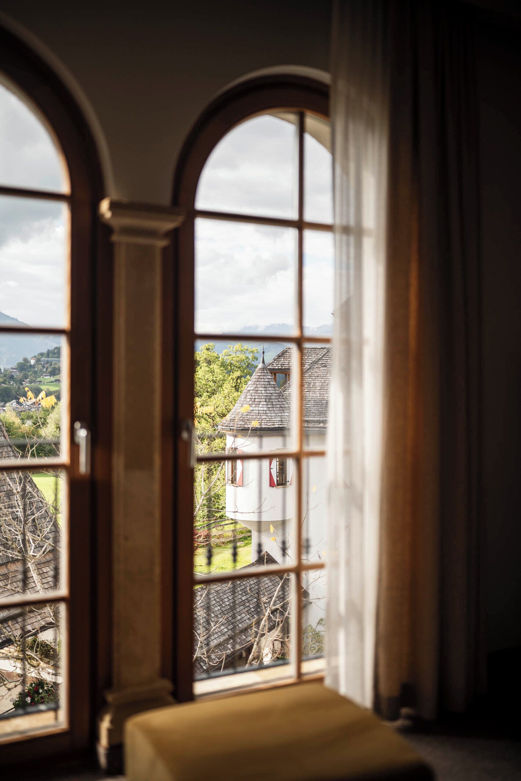 A room with a large arched window offering a view of a village with traditional buildings and a hill in the distance.