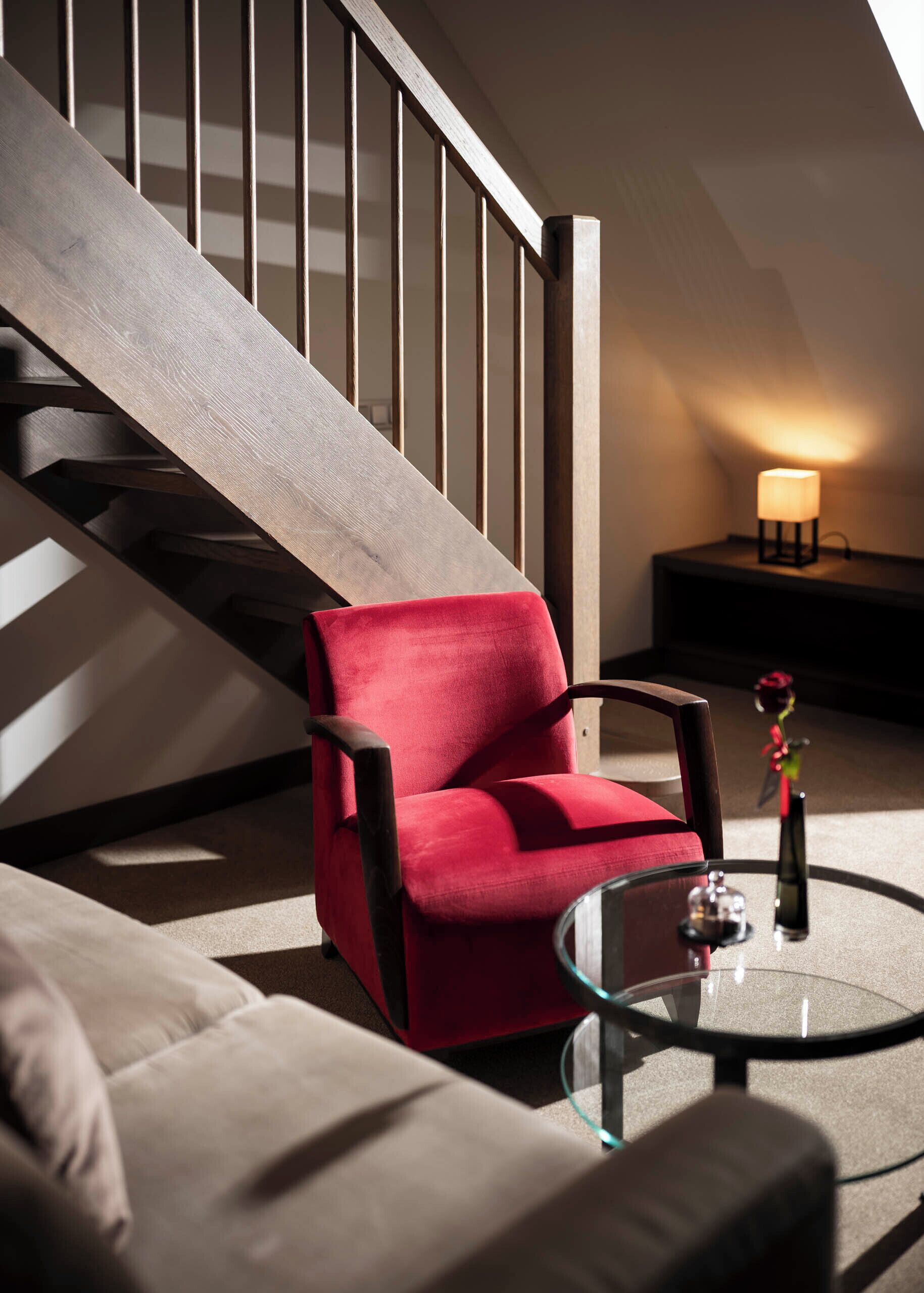 A modern living room with a red armchair, a glass coffee table, and a staircase with a wooden railing.