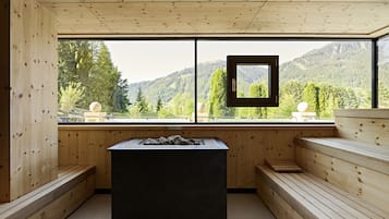 A sauna room with wooden benches and a central table, large windows offering a view of a forested mountain.