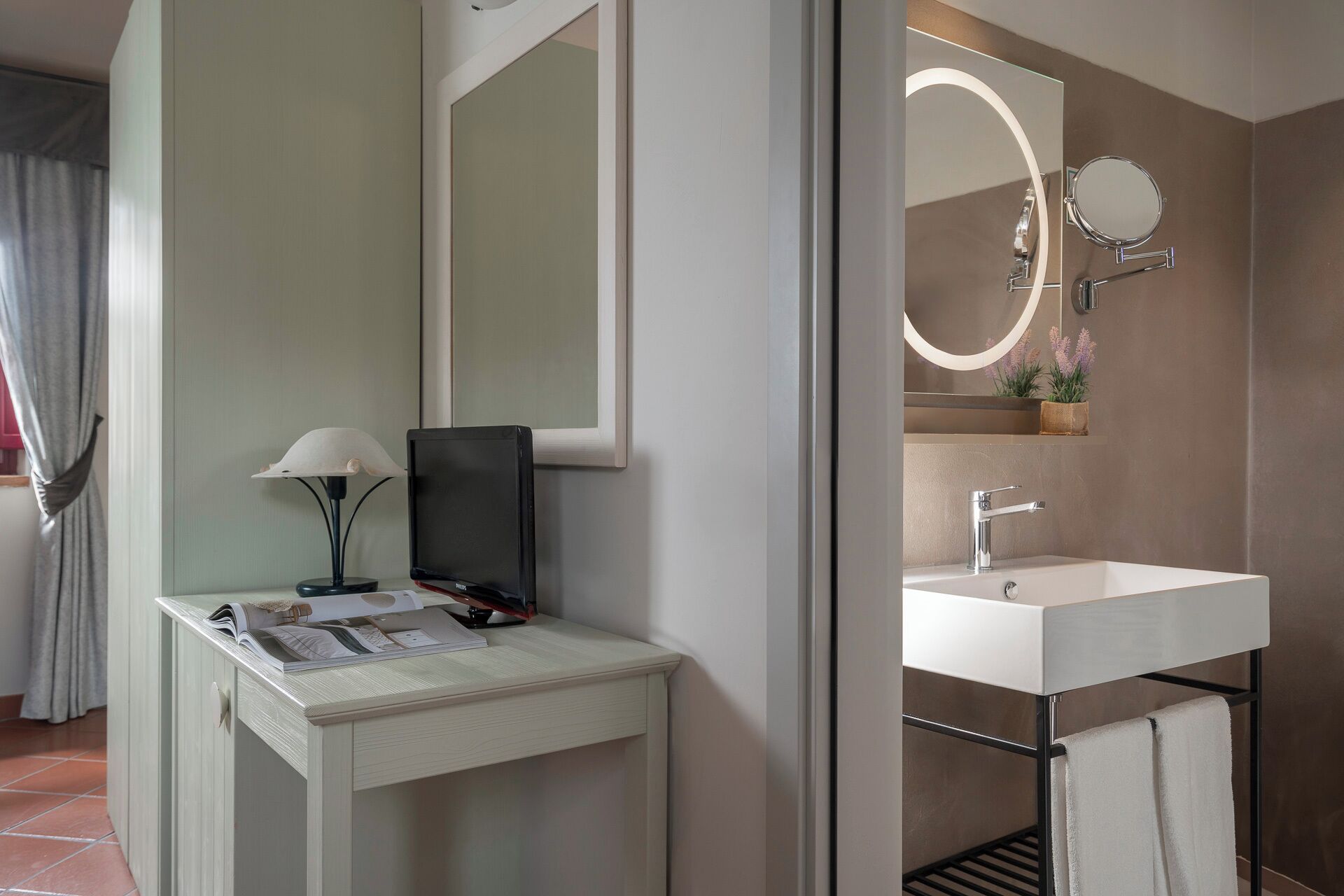 Ein Badezimmer mit weißem Waschbecken, einem beleuchteten Spiegel und einem kleinen Tisch mit Lampe und Buch.