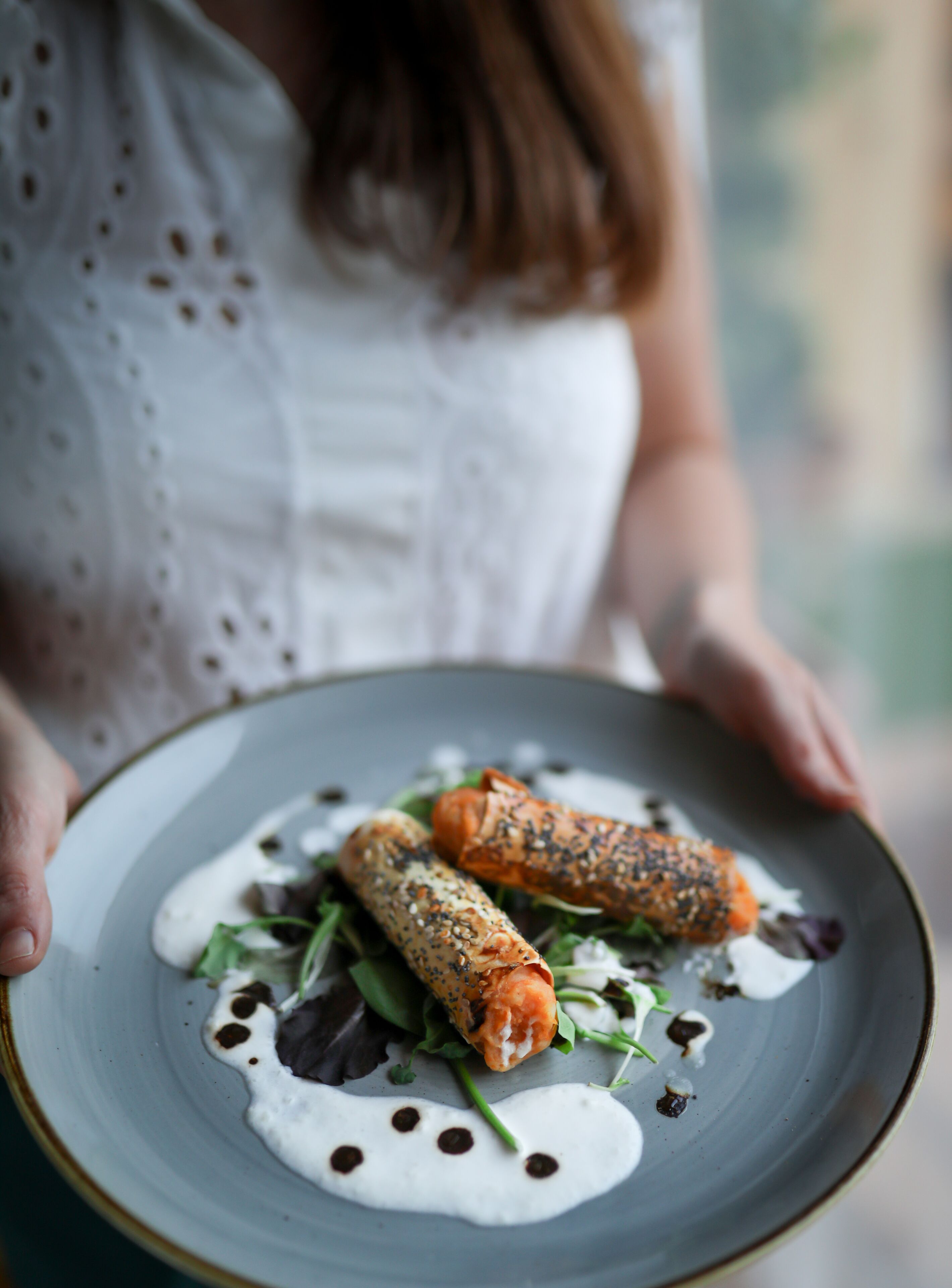Una persona sosteniendo un plato con dos porciones enrolladas, posiblemente comida, adornado con vegetales verdes y salsa blanca.