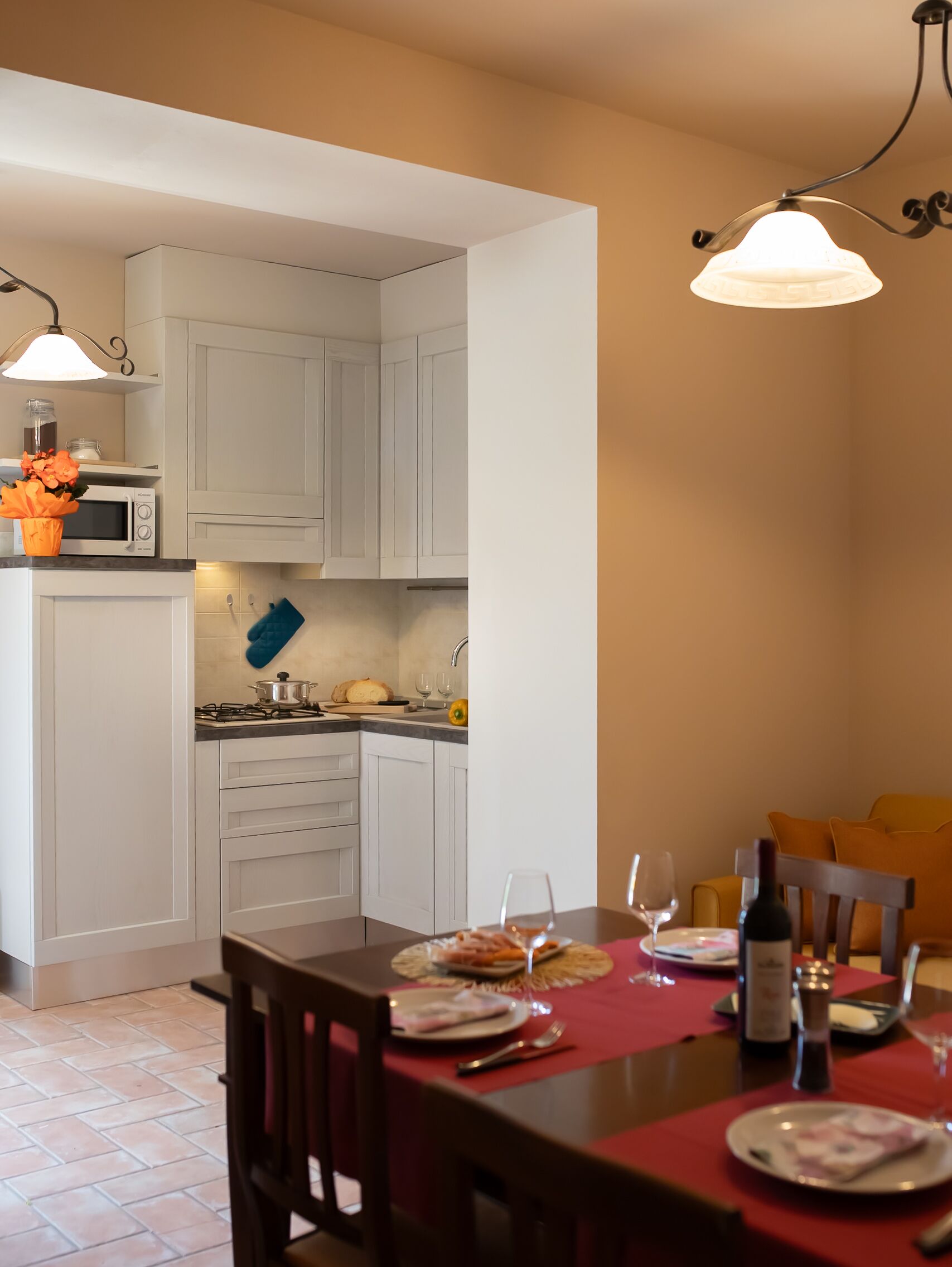 Una cocina moderna con armarios blancos, un microondas y una estufa a gas. Hay una mesa de comedor preparada con platos, vasos y una botella.