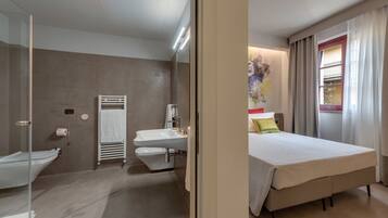 Habitación de hotel moderna con cabina de ducha de vidrio, lavabo doble, cama con cubrecama blanco y ventana con cortinas.