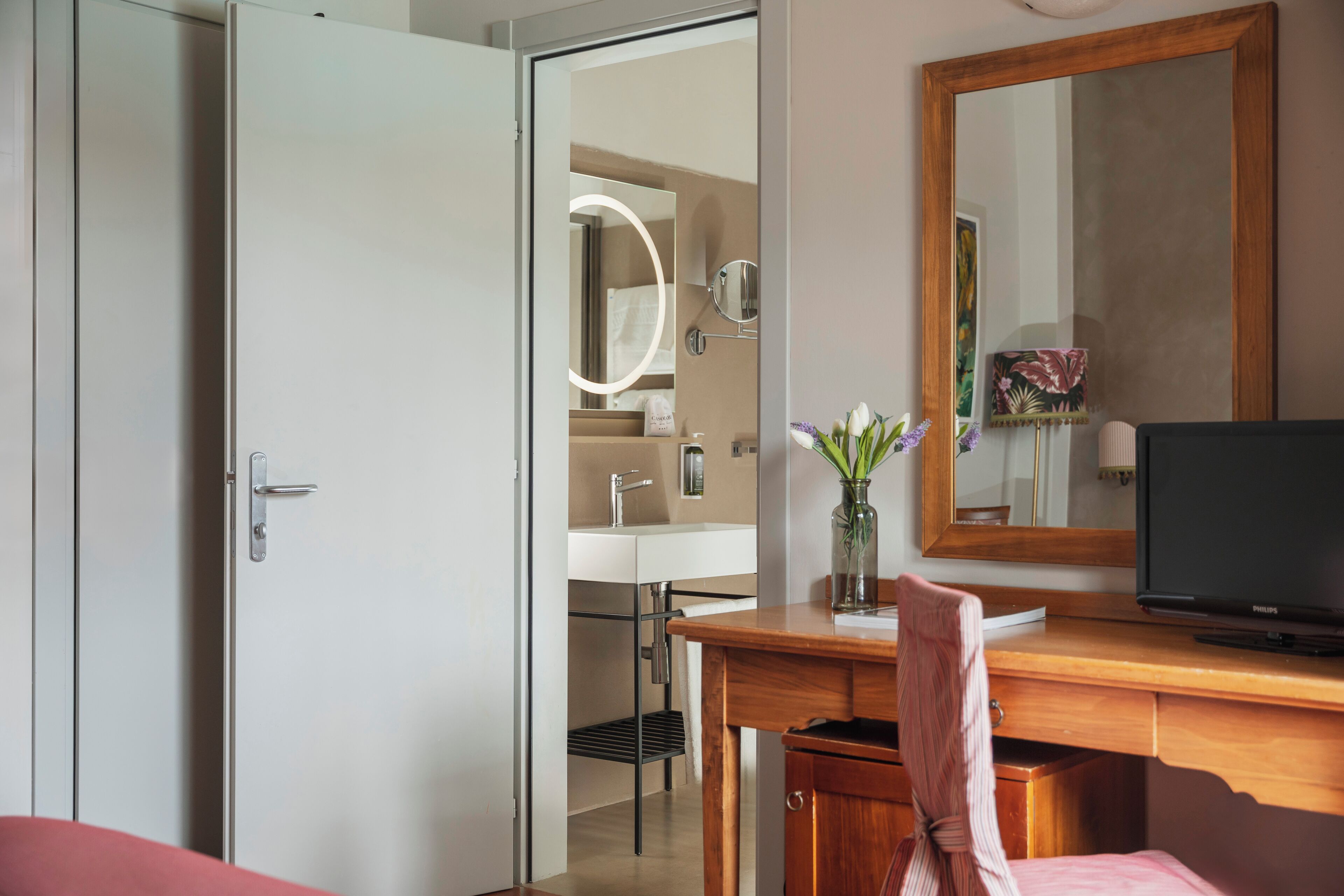 A hotel room with a wooden desk, a television, a mirror, and a bathroom visible through an open door.