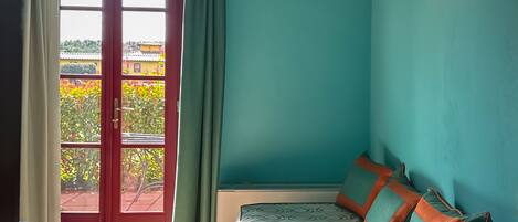 Una habitación con cama, ventana con cortinas, lámpara fijada a la pared y vistas al exterior con vegetación.