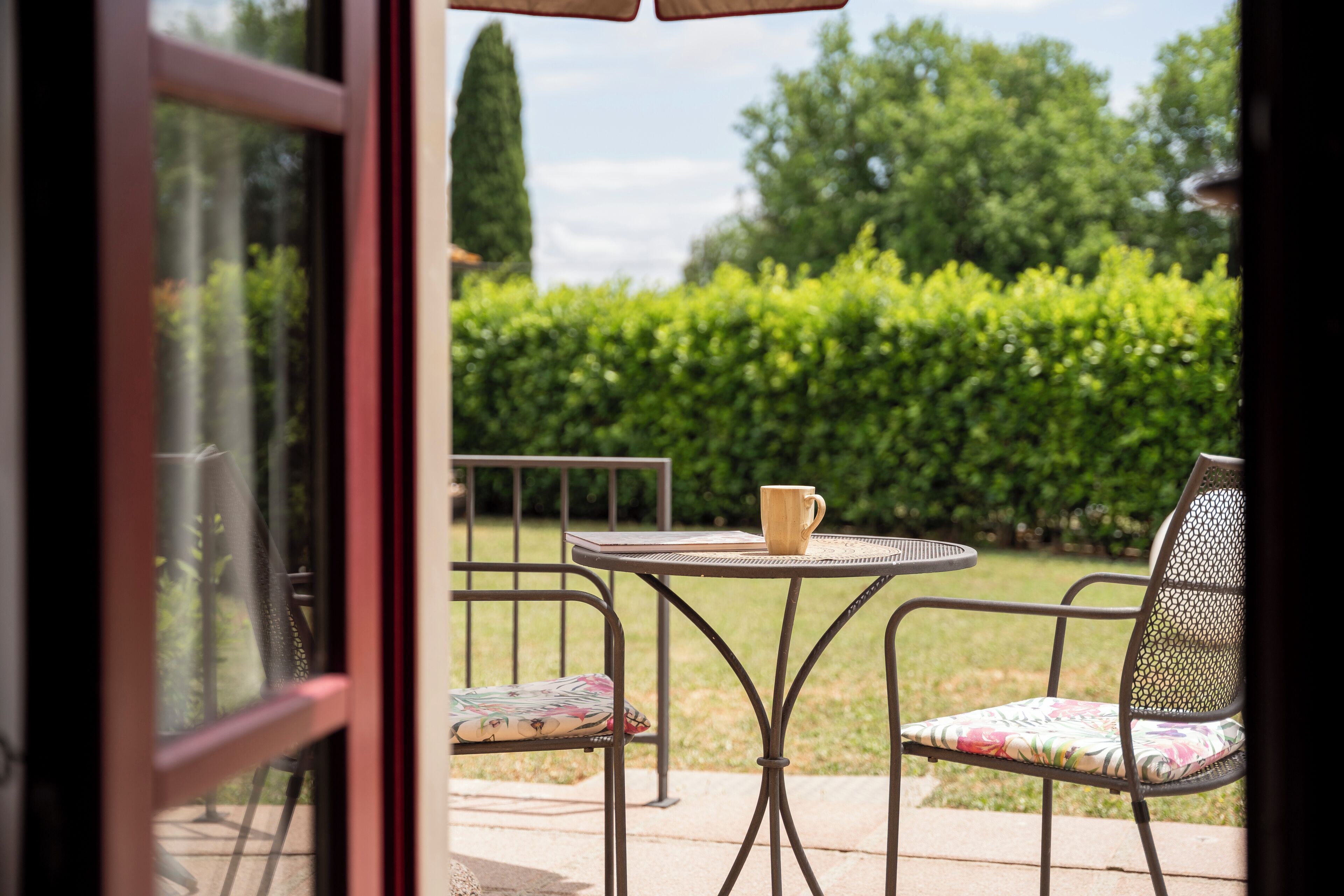 Een terras buiten met een ronde metalen tafel en twee stoelen, waarvan er één een bloemenkussen heeft.