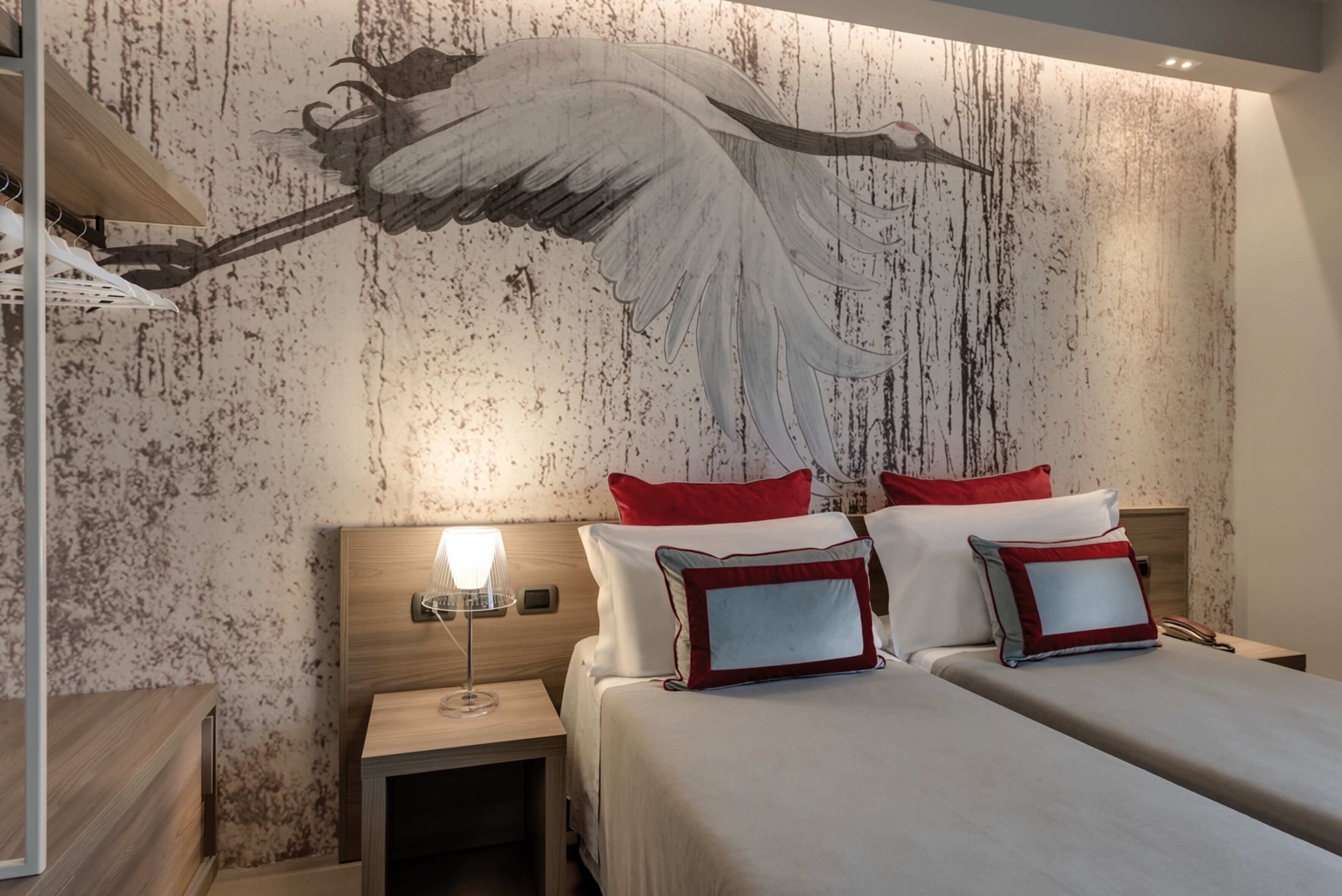 A hotel room with a large mural of an angel on the wall, a wooden headboard, a bedside table with a lamp, and a bed with red and white bedding.