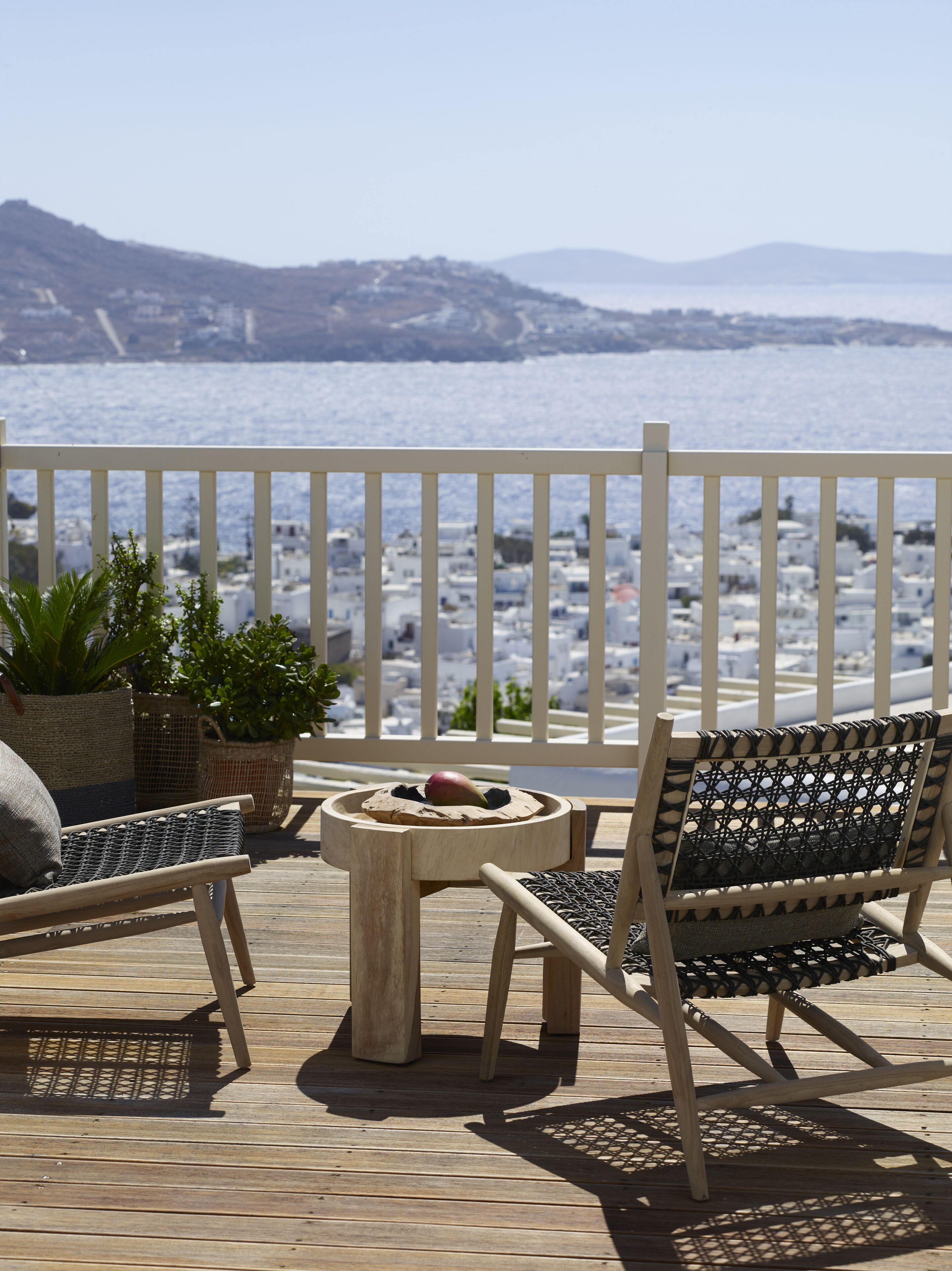 Una terraza de madera con dos sillas de mimbre y una pequeña mesa redonda, con vistas a la costa donde se ven edificios blancos y una valla blanca.