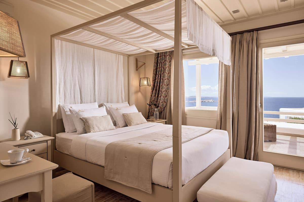 Een hemelbed met witte lakens, een nachtkastje met lamp, en uitzicht op zee door grote ramen.